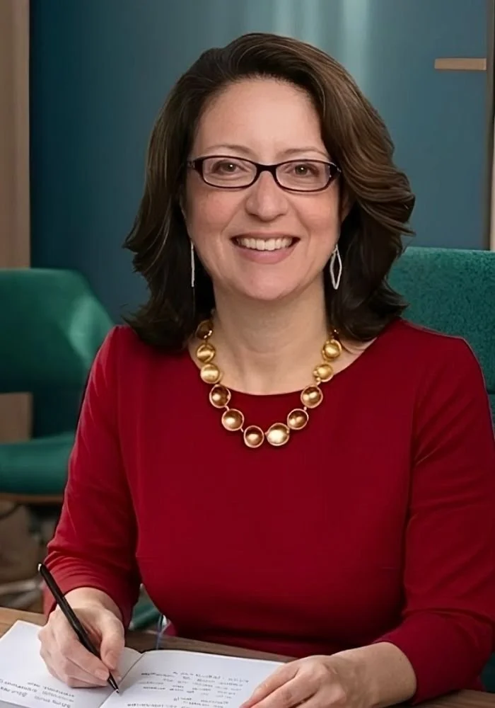 Wendy Maldonado wearing a red dress with a pearl necklace, smiling while sitting at a desk and holding a pen over an open notebook.