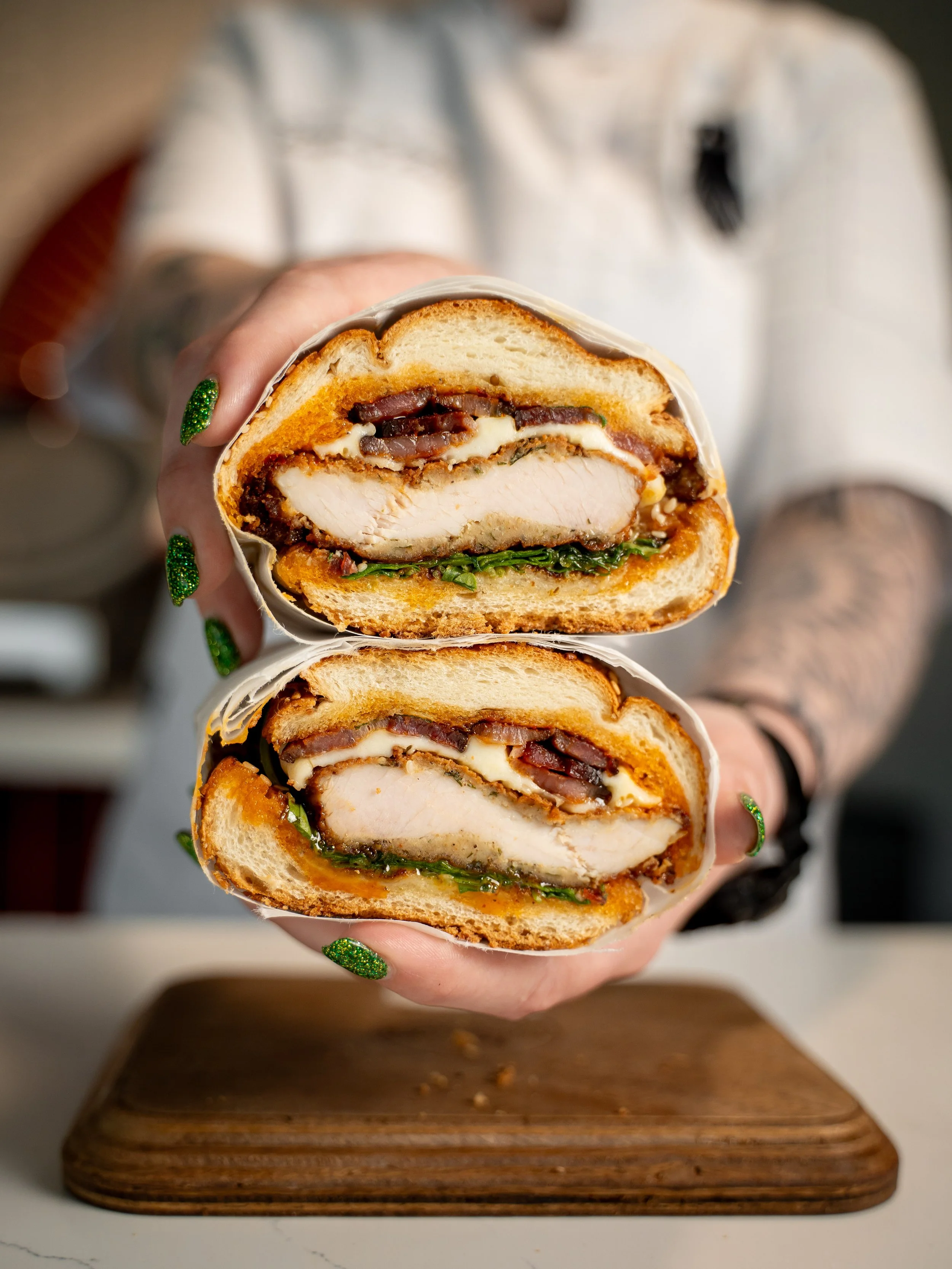 Close-up of a sliced multi-layered sandwich with breaded chicken, bacon, cheese, lettuce, and condiments on a wooden cutting board.