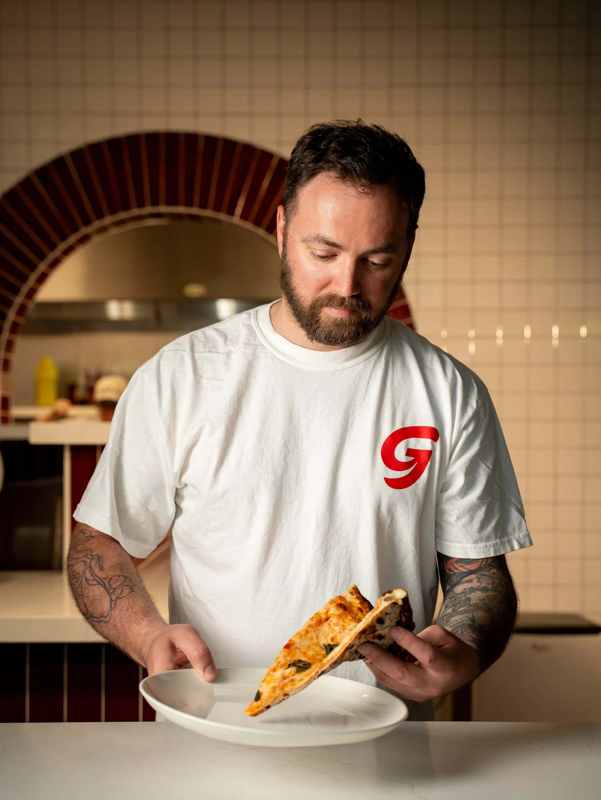 A man with a beard and tattoos on his arms is holding a slice of pizza over a white plate, standing in a kitchen with beige tiled walls and a curved red brick oven in the background.
