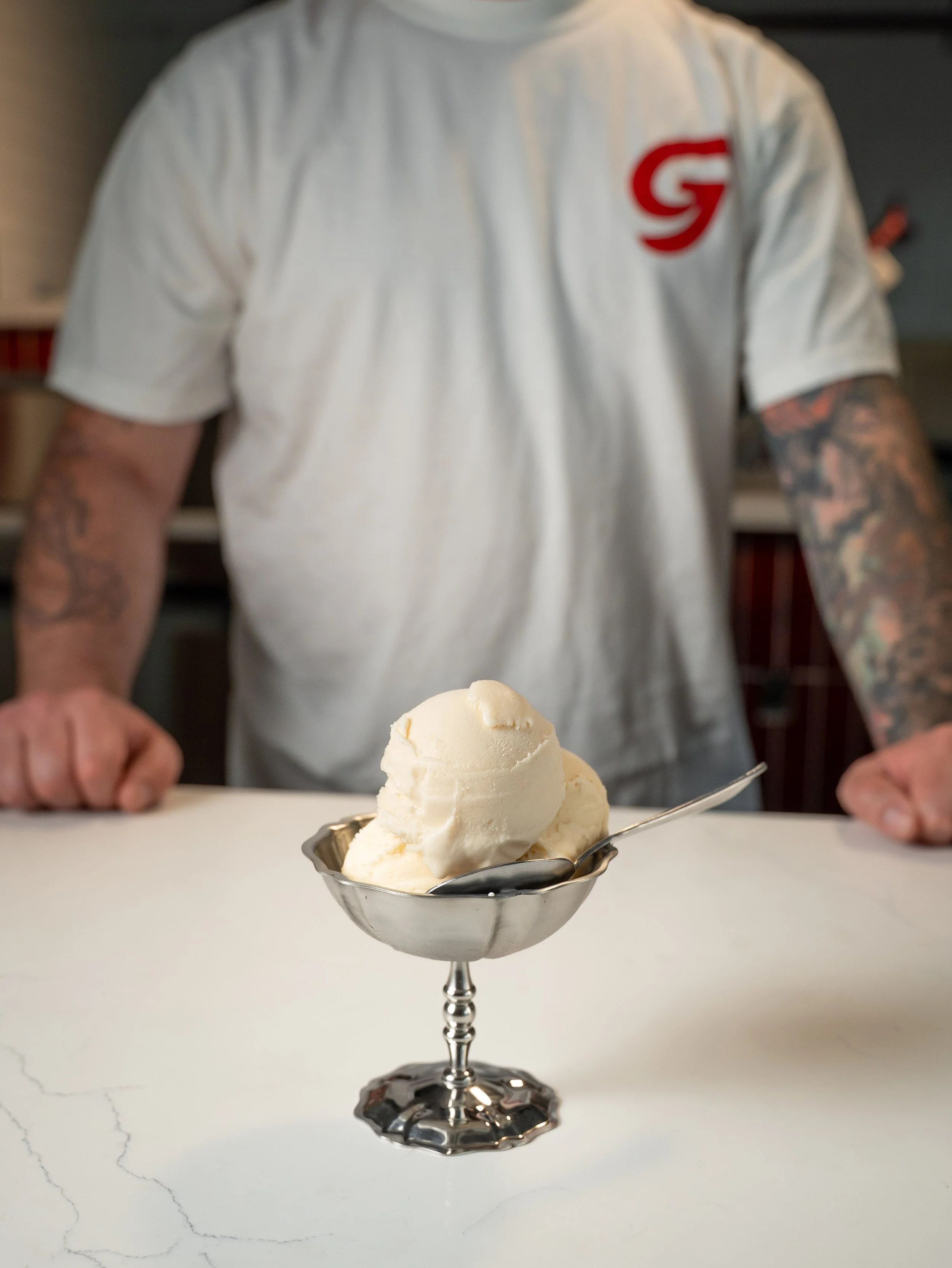 A person with tattoos on their arms wearing a white t-shirt with a red logo, standing behind a table with a silver ice cream dish containing a scoop of vanilla ice cream in the foreground.
