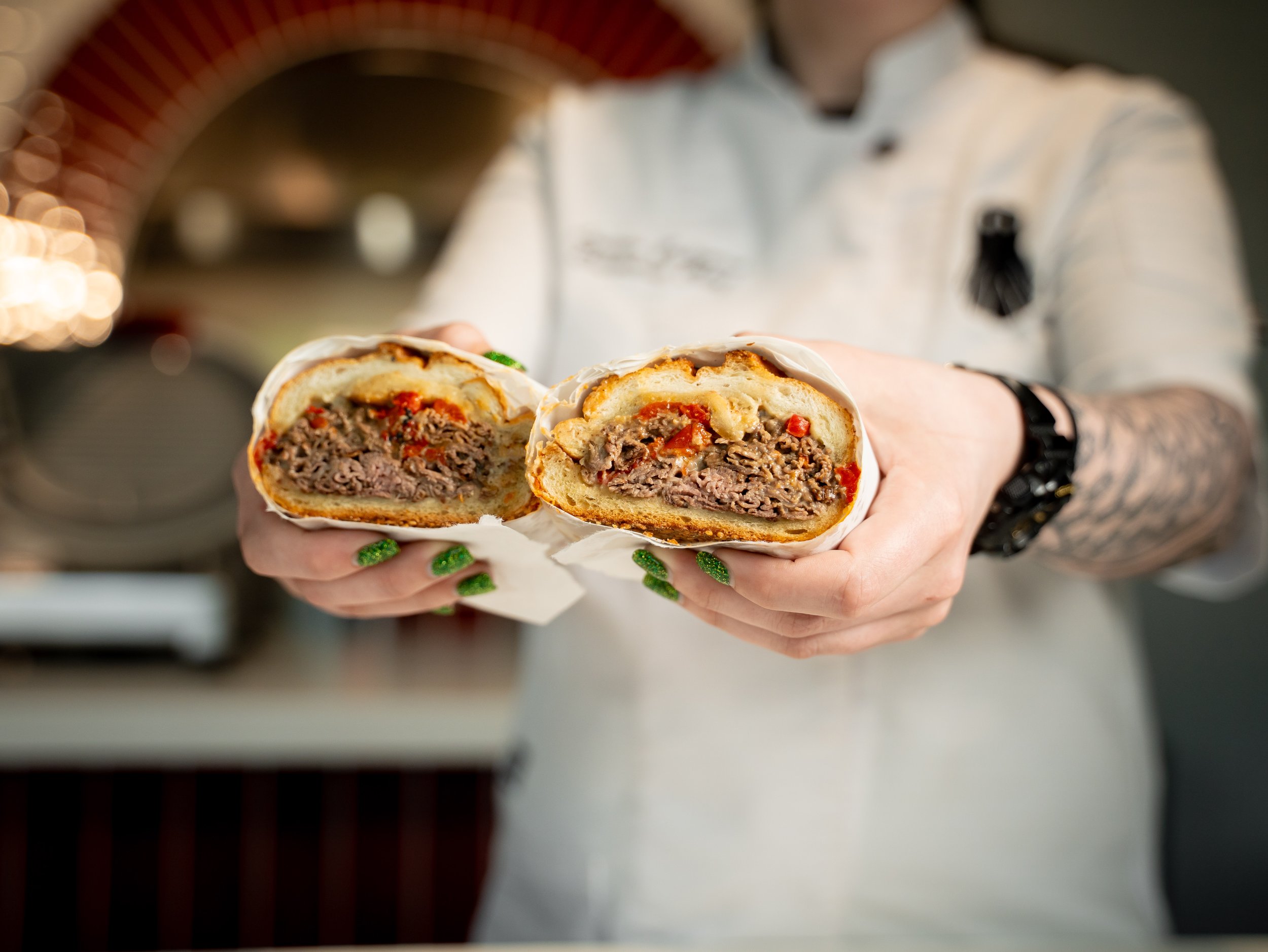 A person holding a sliced beef and vegetable wrap or burrito, showing the filling inside, with the person wearing a white shirt and having green painted nails.