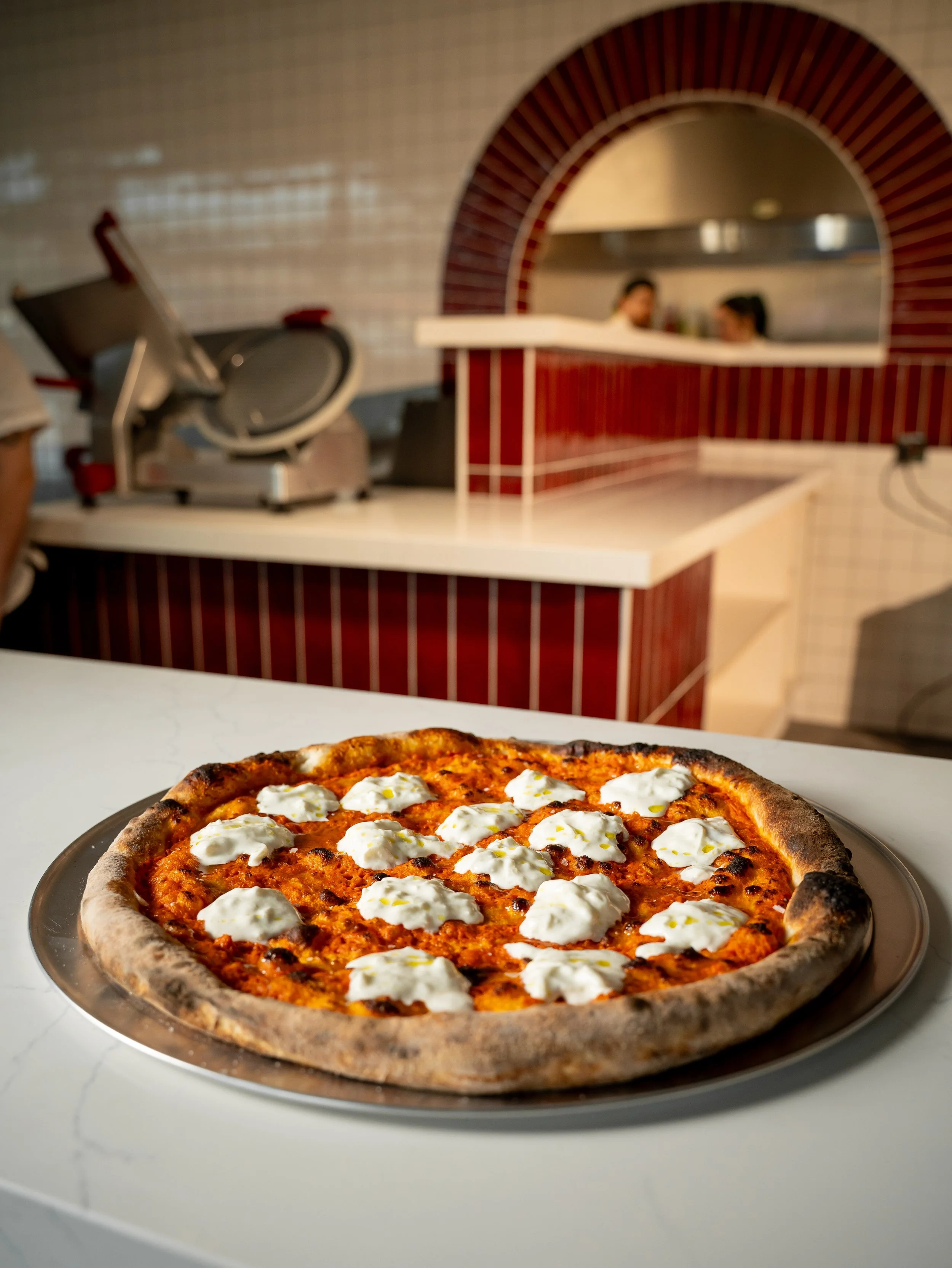 A pizza with a thin crust, tomato sauce, melted cheese, and dollops of ricotta cheese on top, sitting on a silver platter on a white countertop.