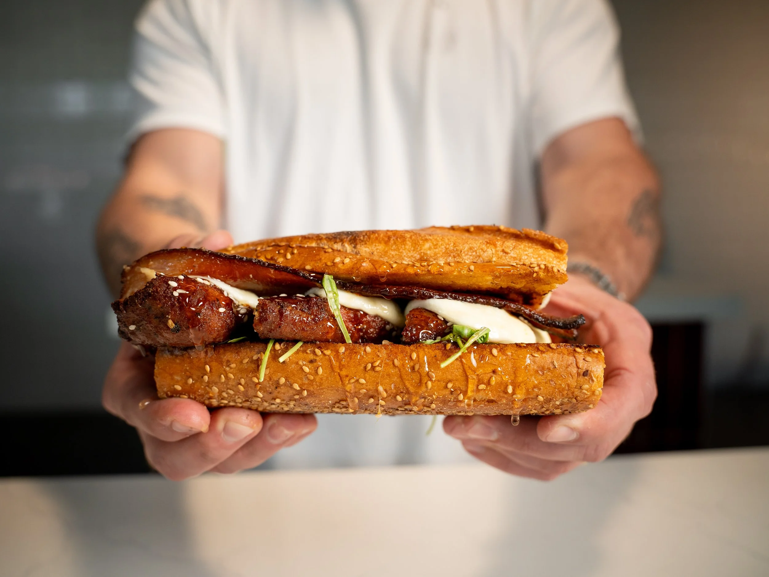 A person holding a sandwich with fried chicken, bacon, cheese, and vegetables in a sesame seed bun.
