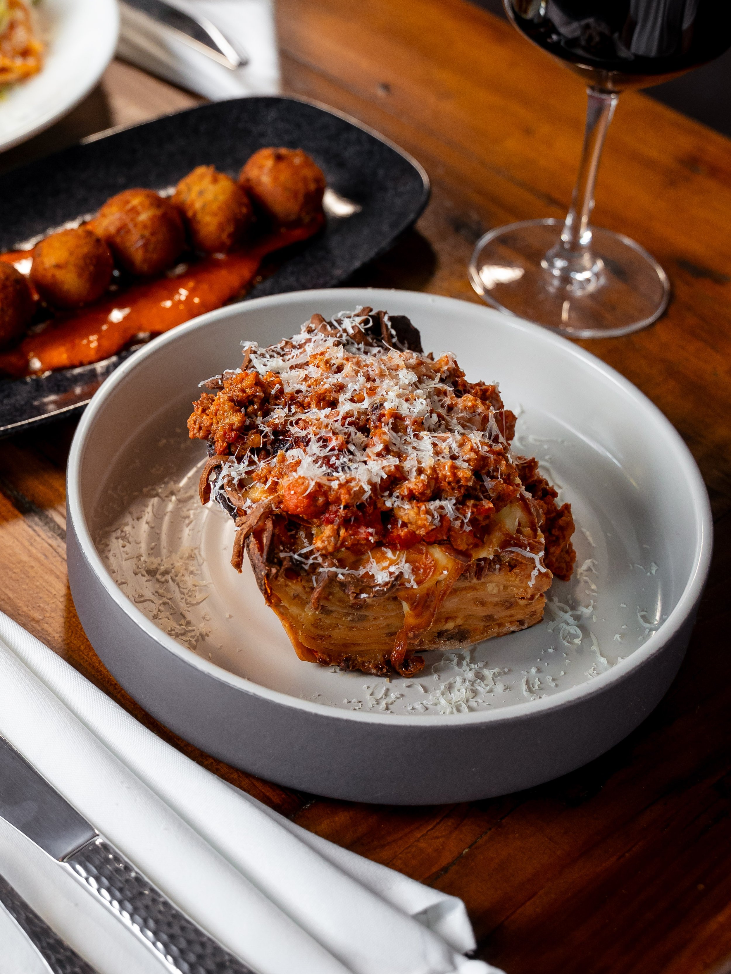 A white bowl with baked pasta topped with cheese, a black plate with lasagna and a black plate with meatballs in tomato sauce, a glass of red wine on a wooden table setting with silverware and a white napkin.