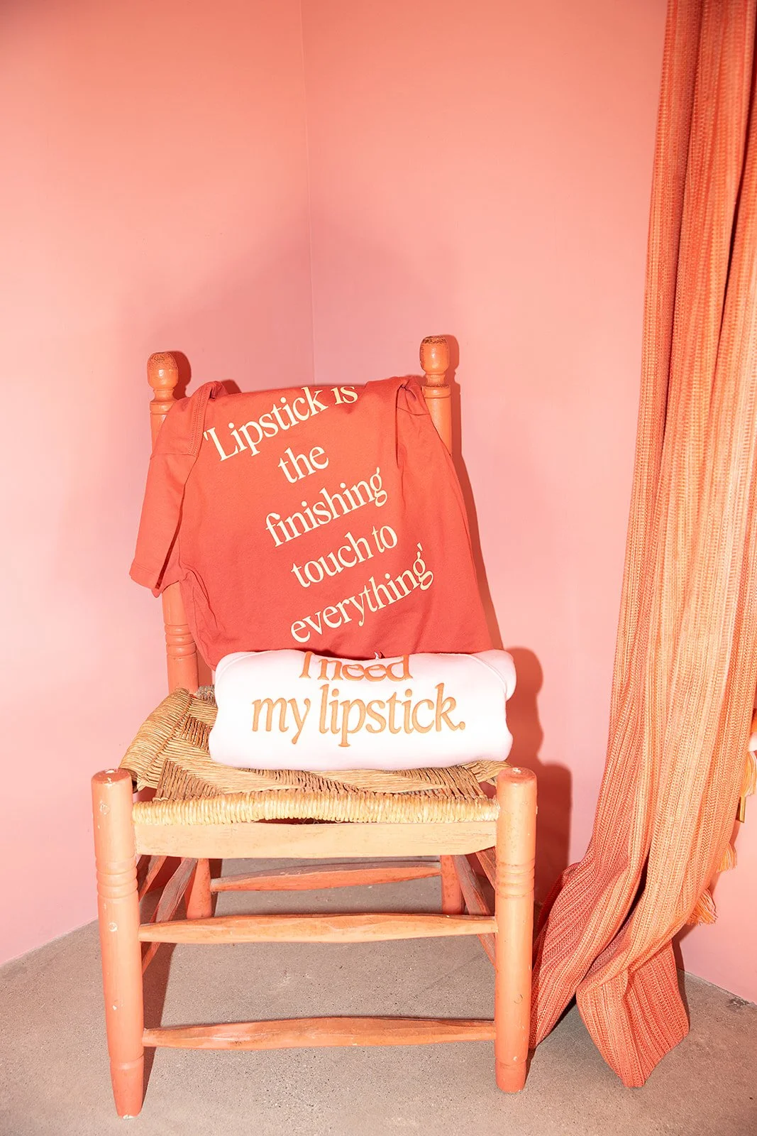 A wooden chair with a woven seat against a pink wall, holding two pillows. The larger pillow is pink with white text that says, "Lipstick is the finishing touch to everything," and the smaller pillow is white with orange text that says, "I need my lipstick." A pink fabric drapes over the back of the chair and a pink curtain hangs on the right side.