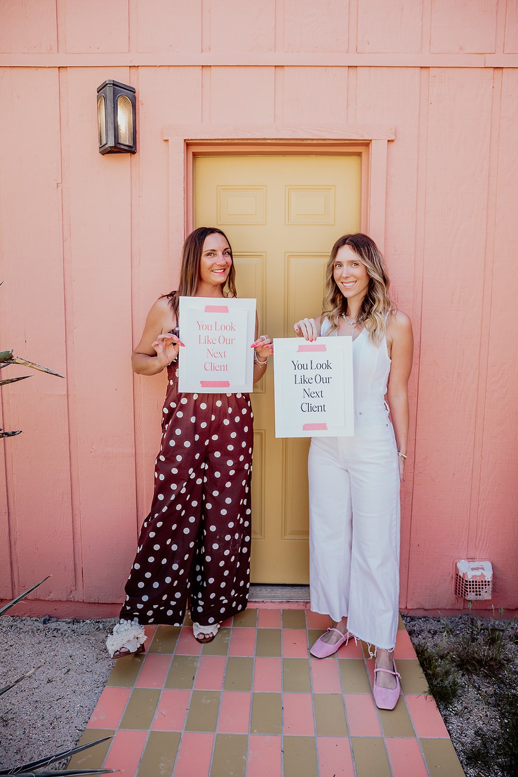 Two women standing in front of a pink wall with a yellow door, holding signs that say "You Look Like Our Next Client."