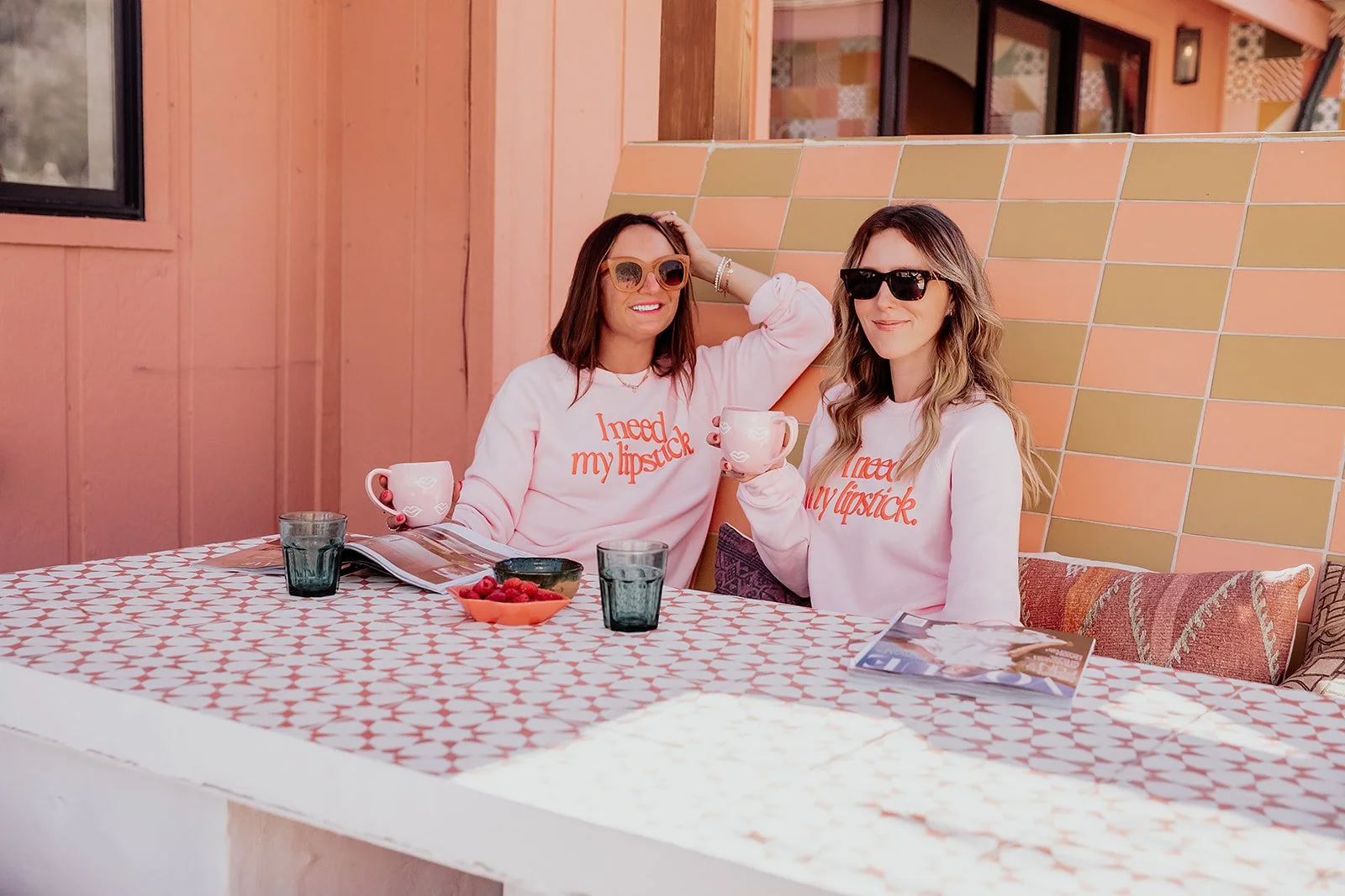 Two women sitting at an outdoor table, wearing matching pink sweatshirts that say "I need my lipstick." They are holding mugs and smiling, wearing sunglasses, with a wall and window in the background, and a magazine and bowls of berries on the table.