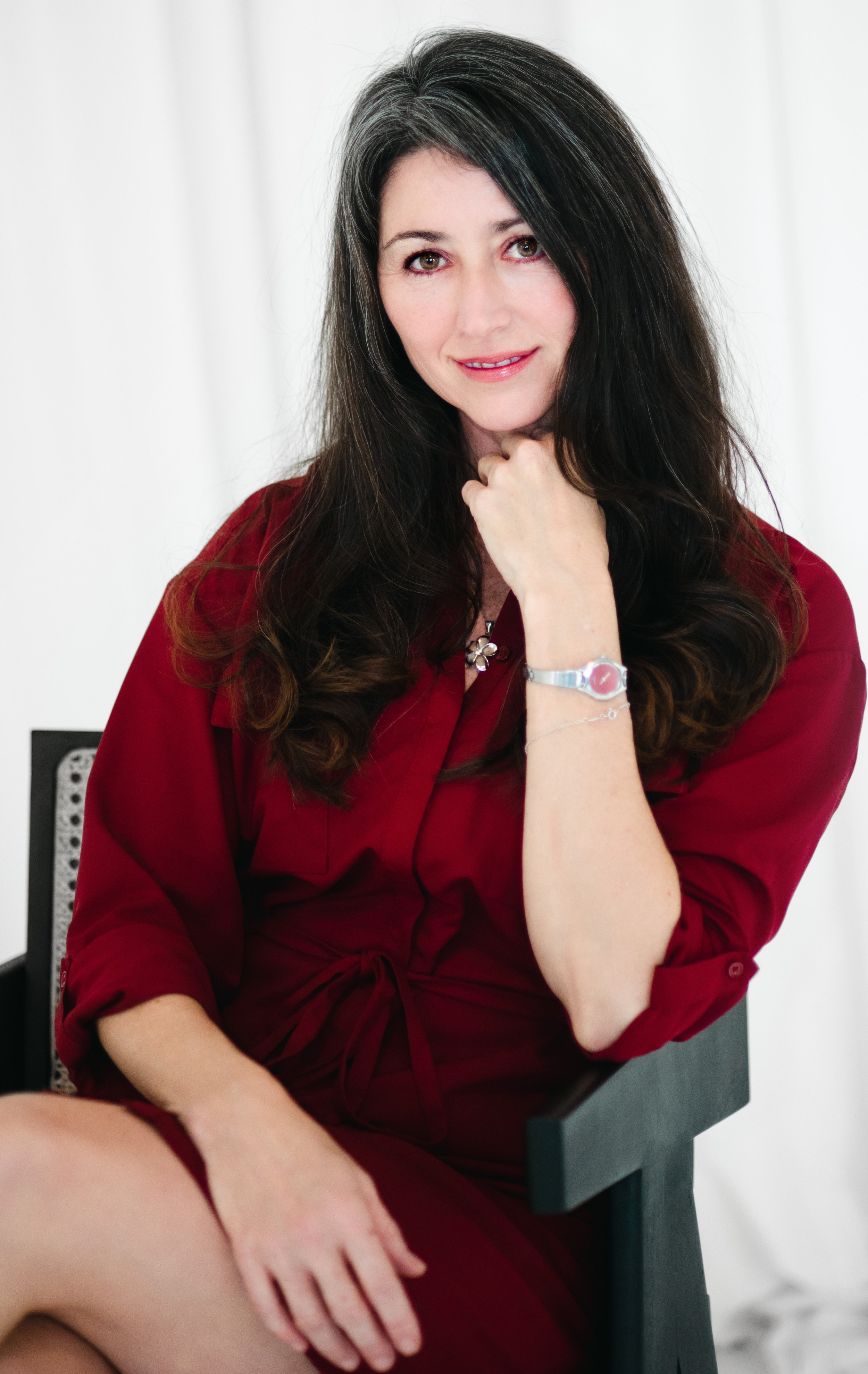 A woman with long dark hair and light skin seated and looking at the camera, wearing a red dress and silver watch, with her head resting on her hand.