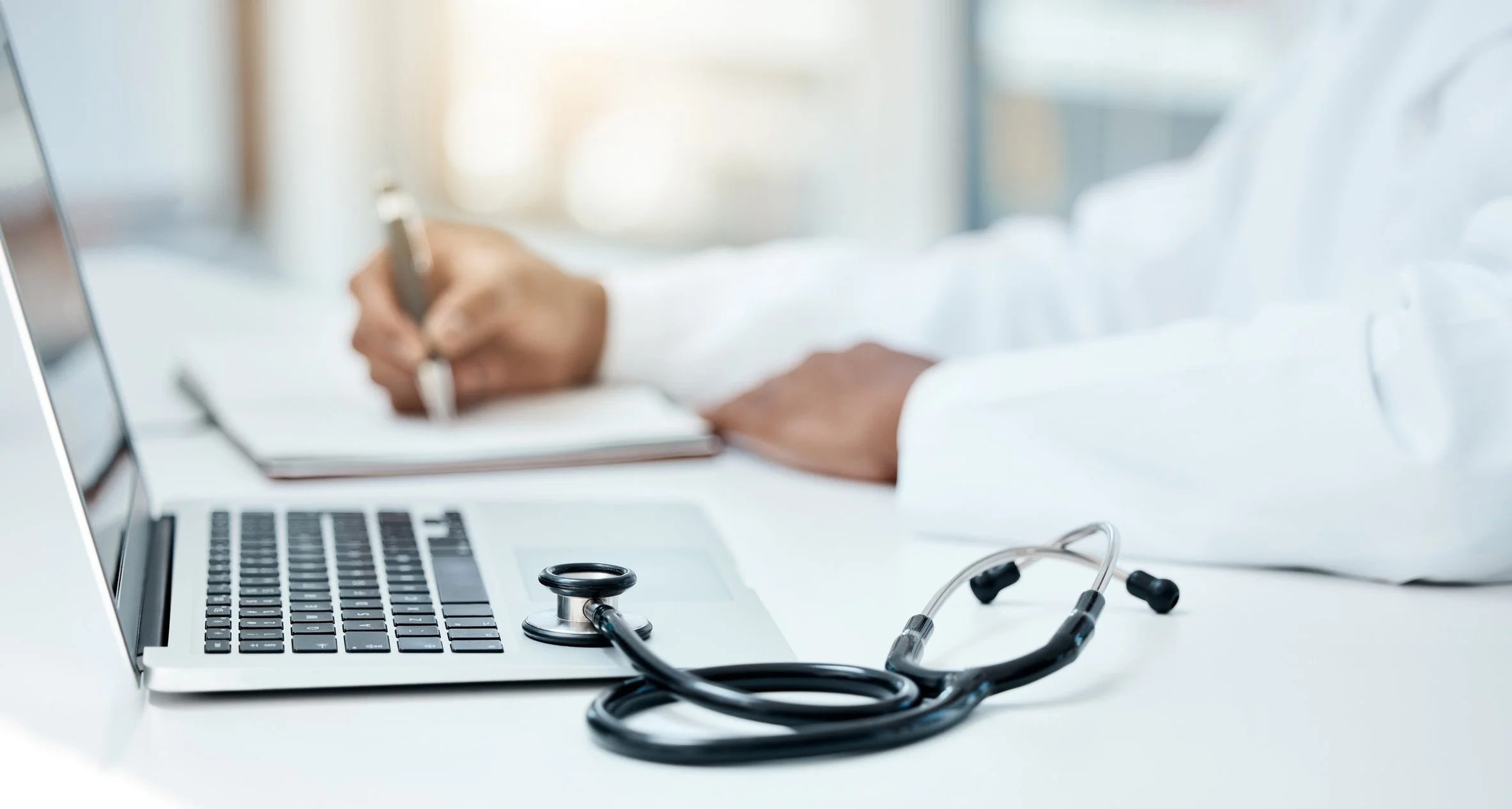 A stethoscope on a laptop keyboard and a doctor writing notes in the background.