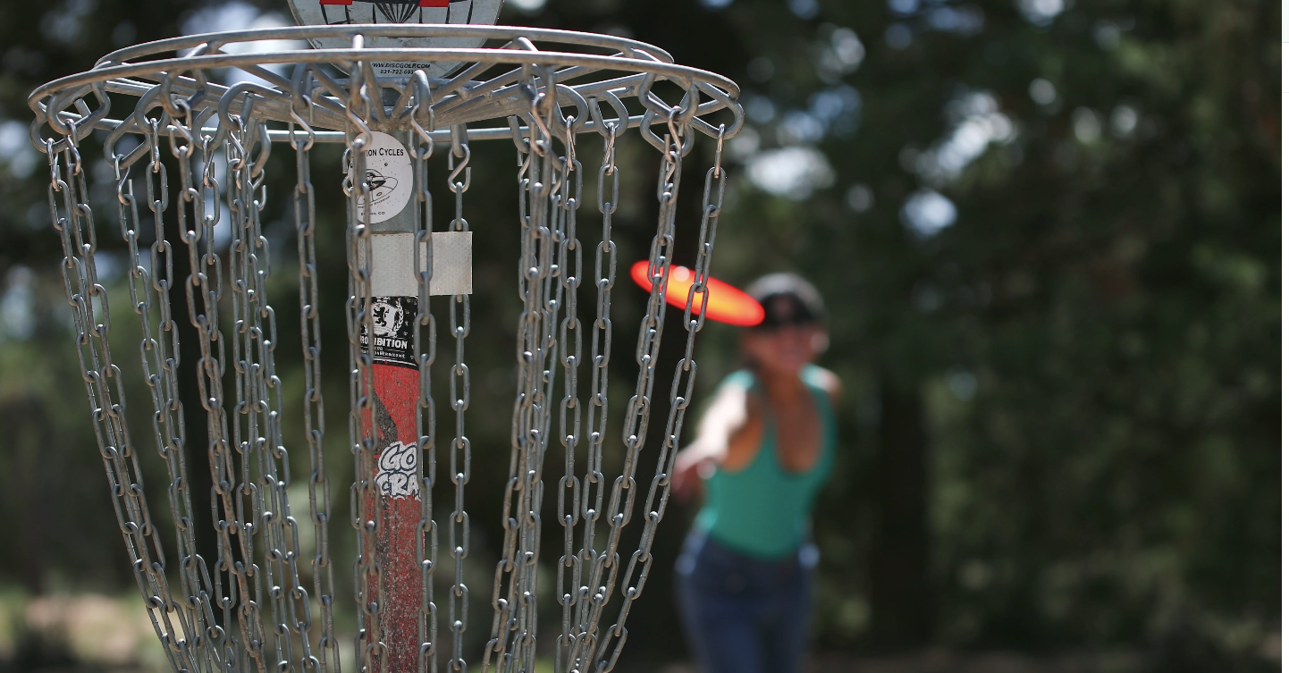 Close-up of a disc golf basket with a person holding a frisbee in the background.
