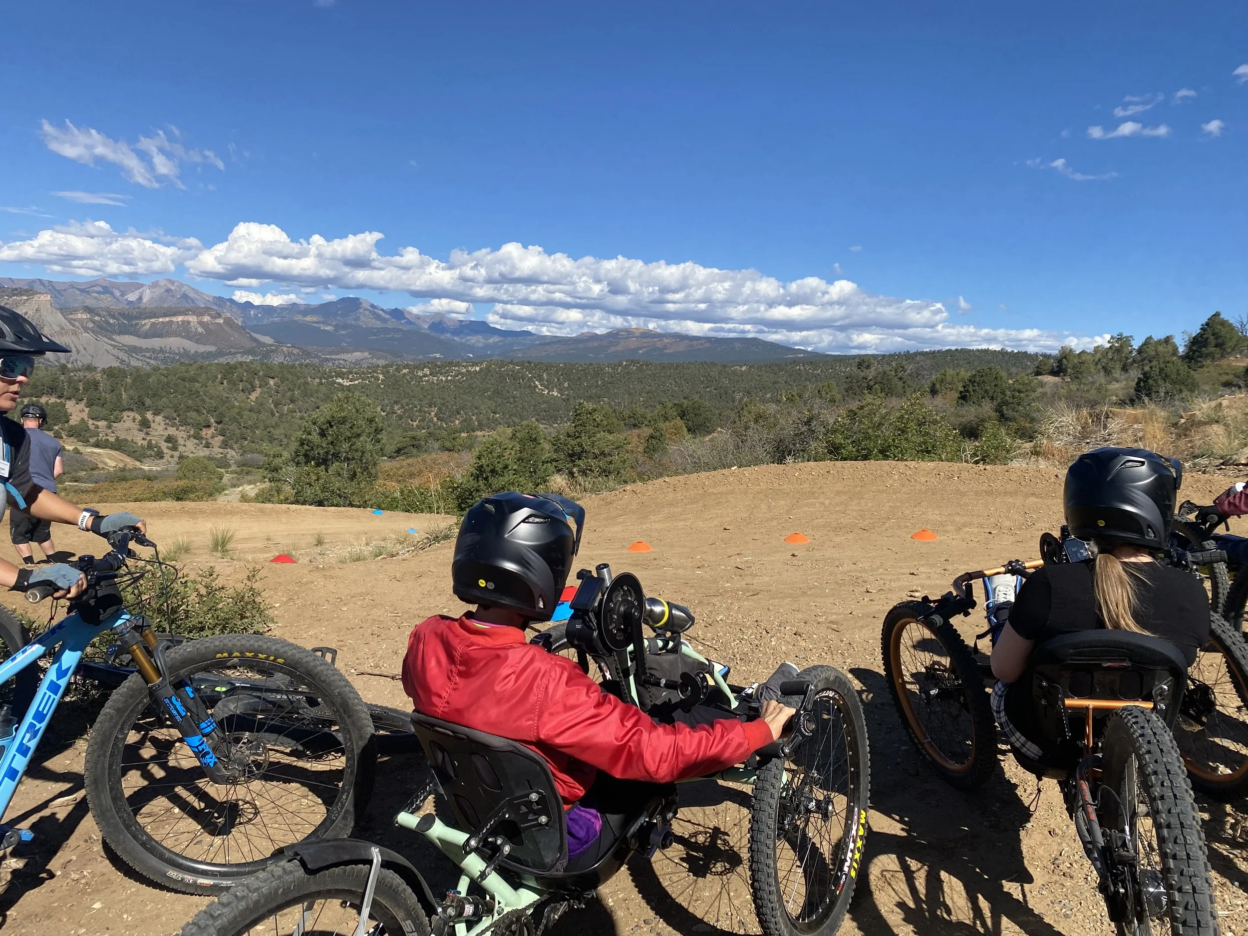 A group of people on mountain bikes in a dirt trail area with a mountainous landscape and blue sky with scattered clouds in the background.