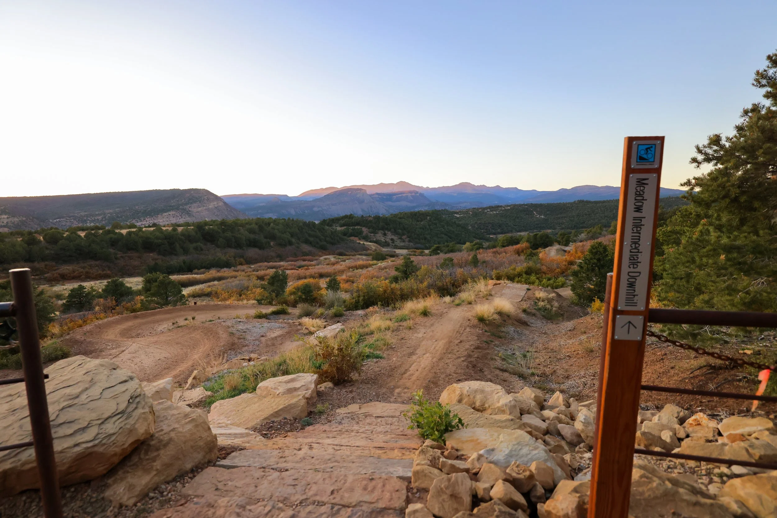 Scenic view of mountains and lush forested landscape with a dirt trail and a trail sign indicating a mountain biking downhill trail, near trees and rocks, during daytime.