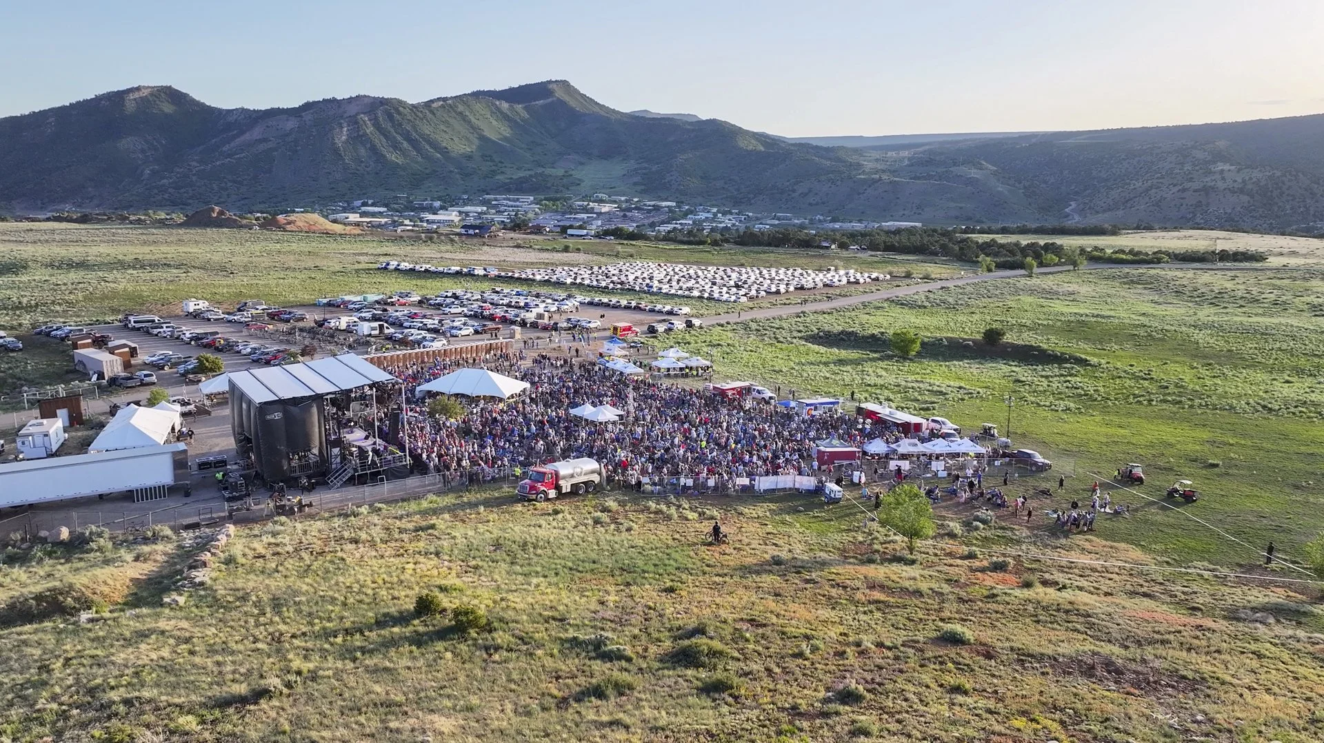 Large outdoor event with a stage and a crowd of people, surrounded by green fields and mountains in the background.