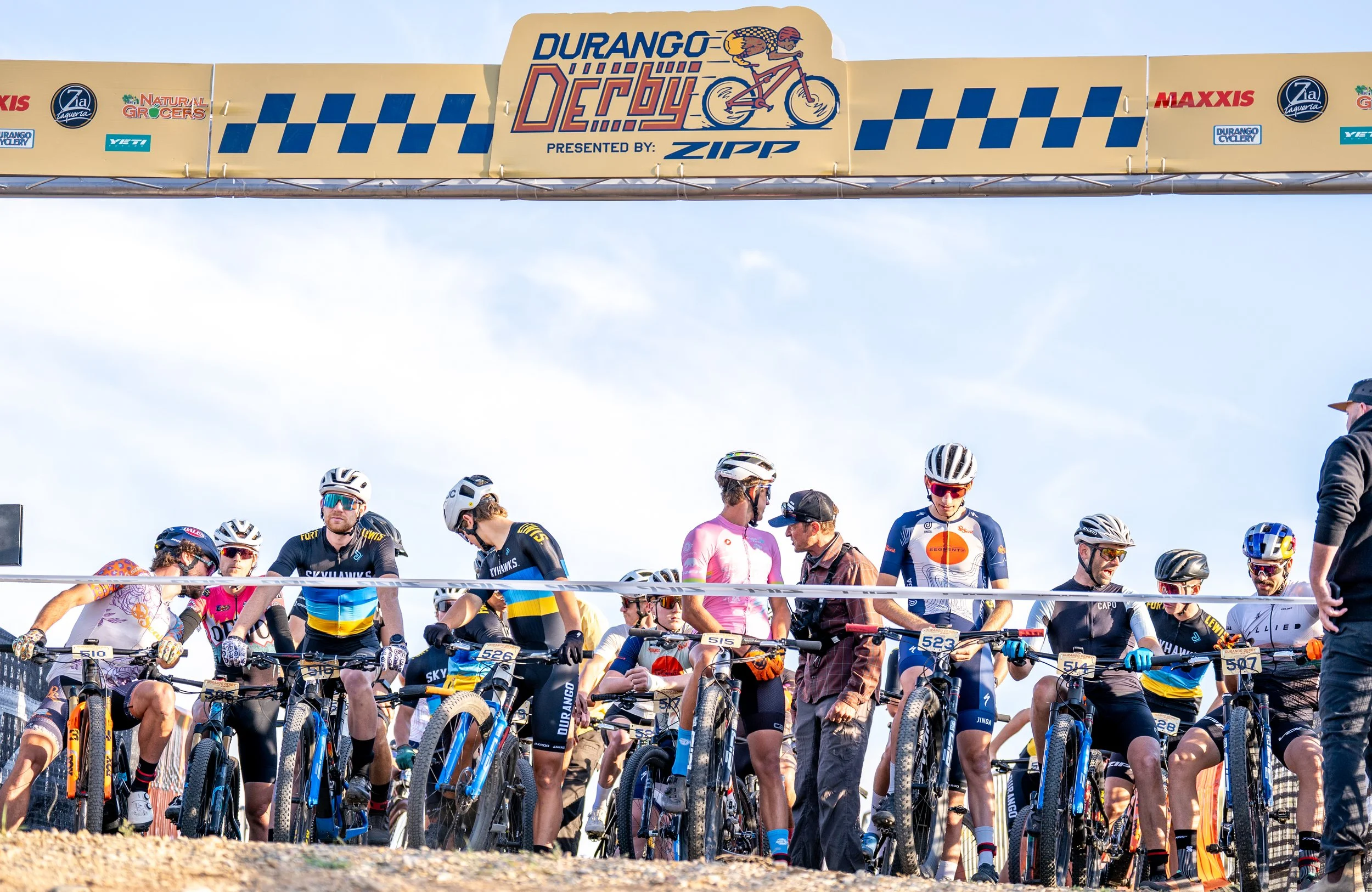 Cyclists gathered at the starting line of the Durango Deby mountain bike race, with a large banner overhead displaying event sponsors and a graphic of a cyclist riding a bike.