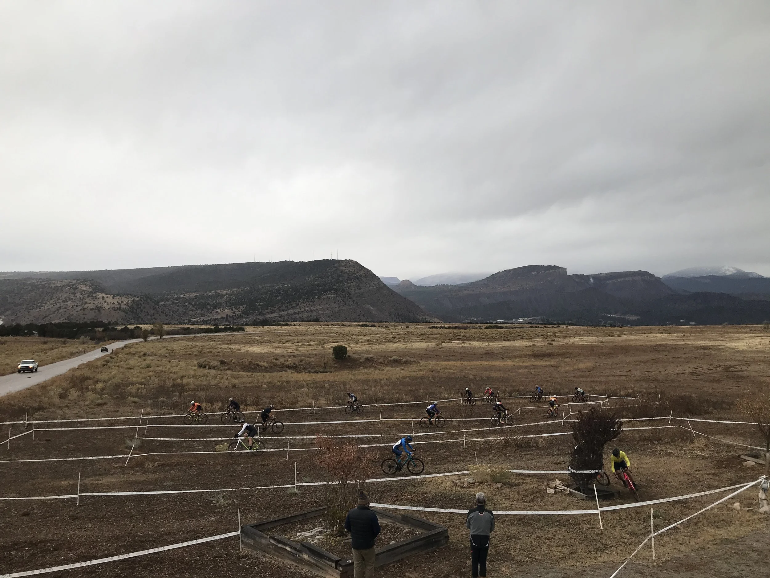 Cyclists participate in a race on a dirt trail bordered by a white fence, set against a backdrop of rolling hills and a cloudy sky.