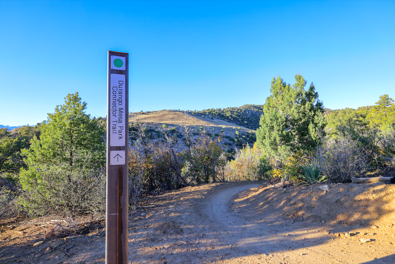 Trail sign indicating the Durangeo Mesa Park Connector Trail, with a dirt trail leading into a landscape of trees and hills under a clear blue sky.
