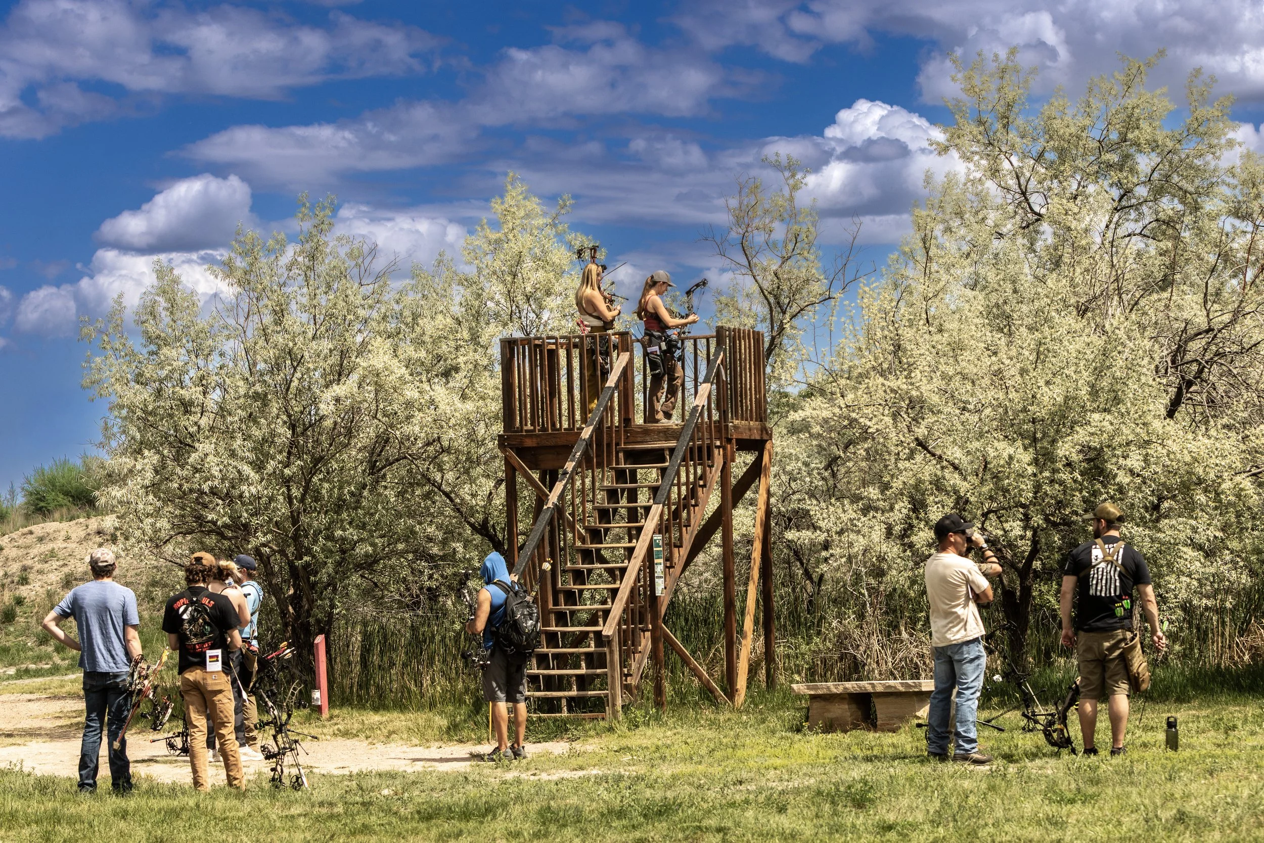 People in outdoor setting near a wooden observation tower with stairs, surrounded by trees and blue sky with clouds.