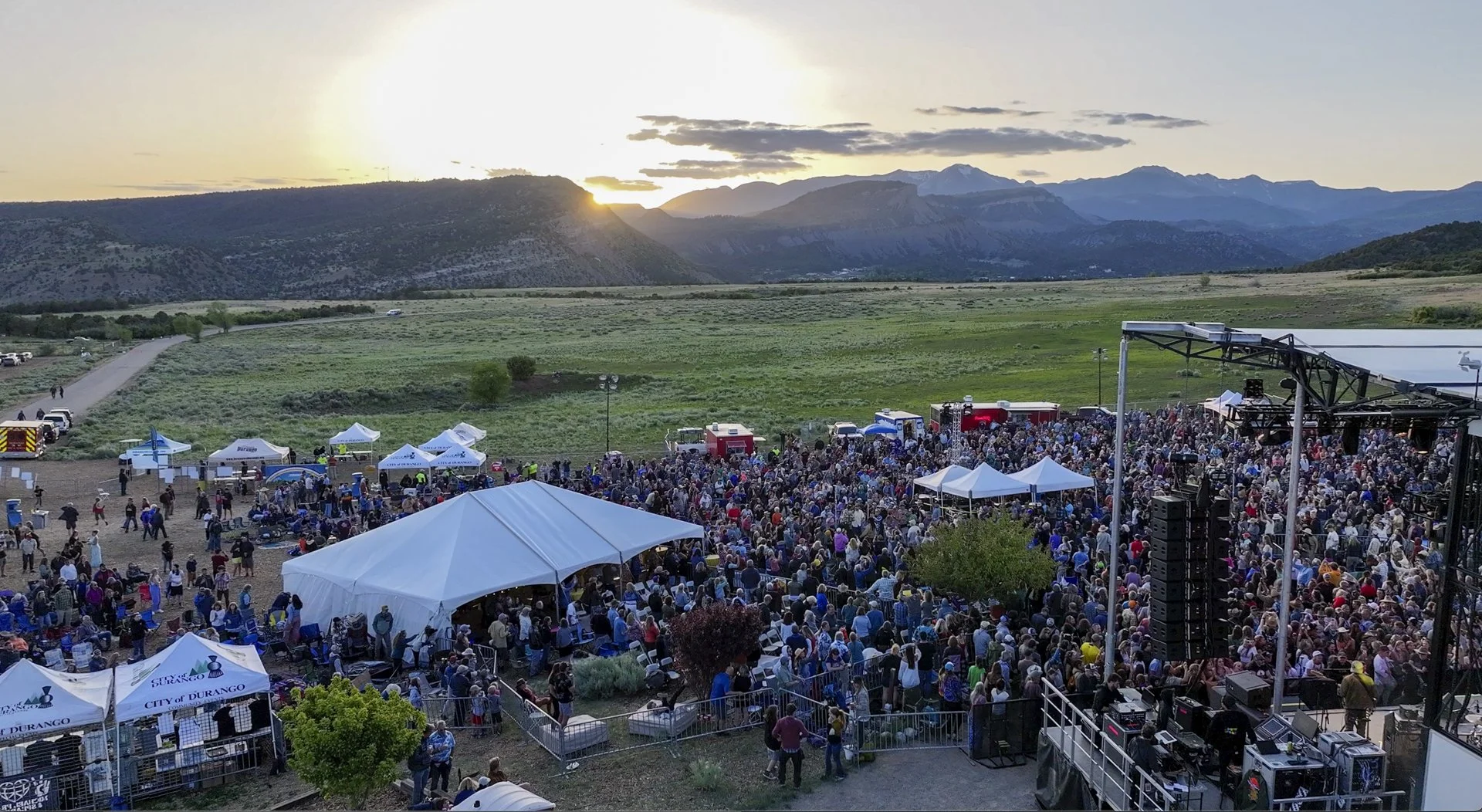 A large outdoor event with a stage and a crowd of people, set against a backdrop of mountains and a setting sun, with tents and food trucks in the area.