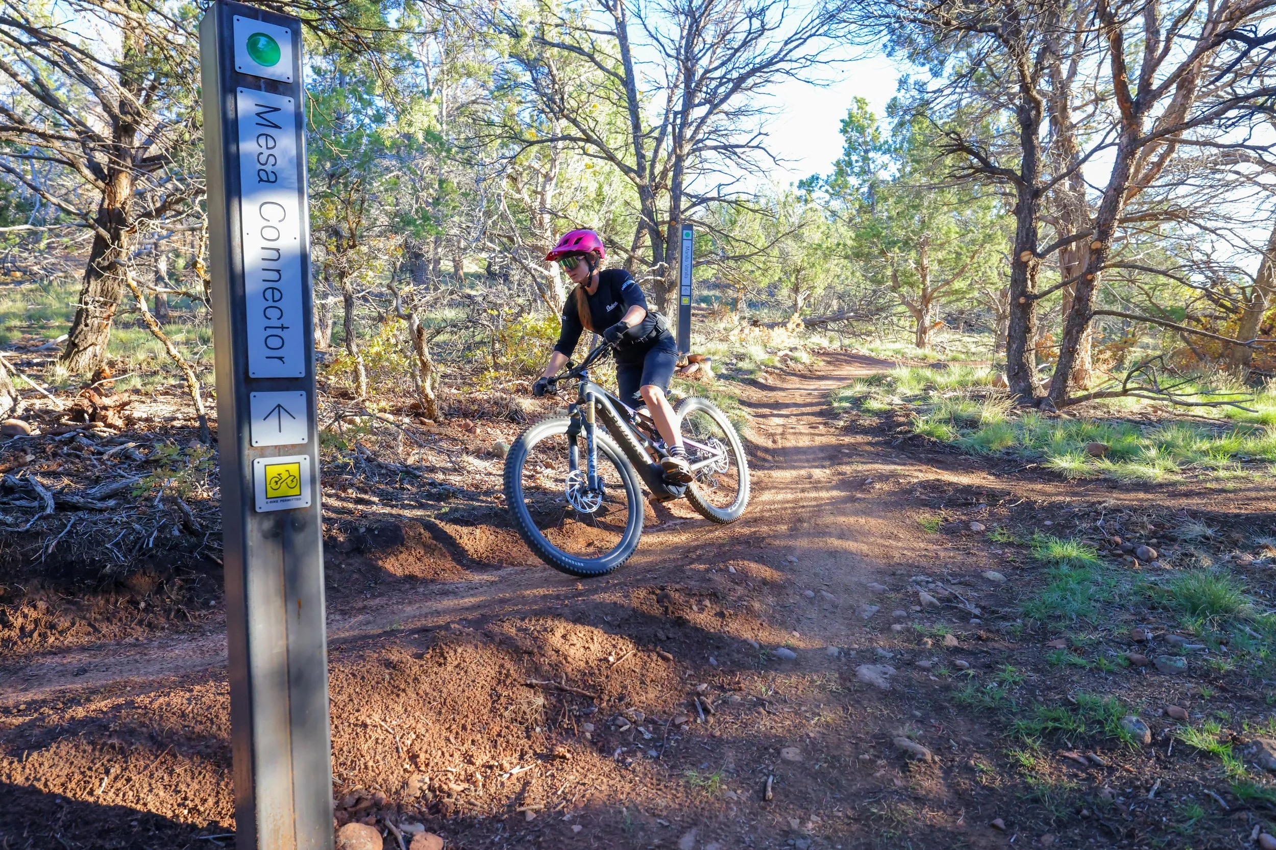 A person wearing a pink helmet, sunglasses, and a black shirt riding a mountain bike on a dirt trail through a forested area with tall trees and green foliage. There are signs on a metal post indicating a trail named 'Messa Connector' and rules for bike riding.