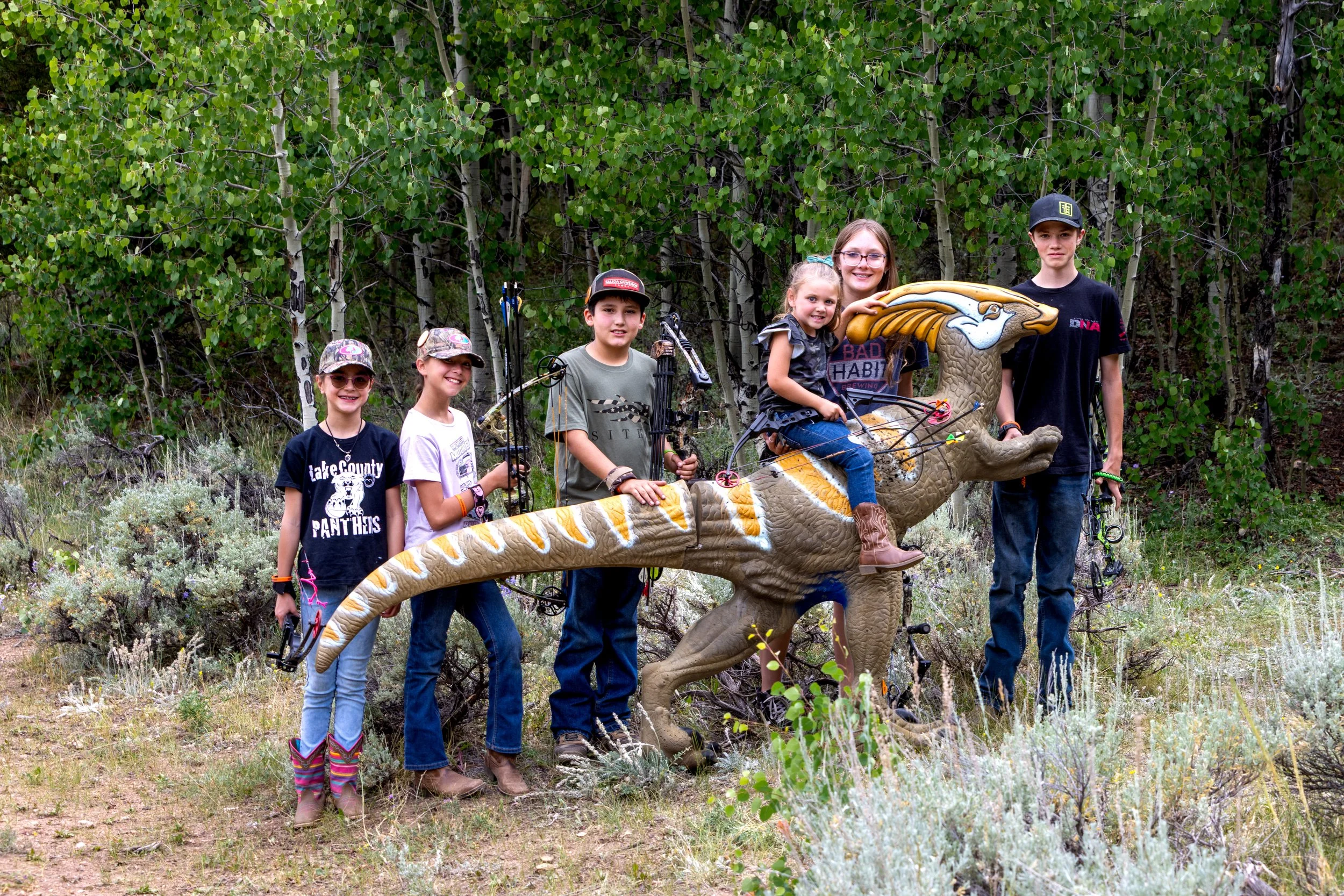 A group of seven children standing outdoors in a forested area. They are posing around a large dinosaur-shaped sculpture that is part of a playground or decorative installation. The children are dressed casually, some with hats, and are holding archery bows and arrows.
