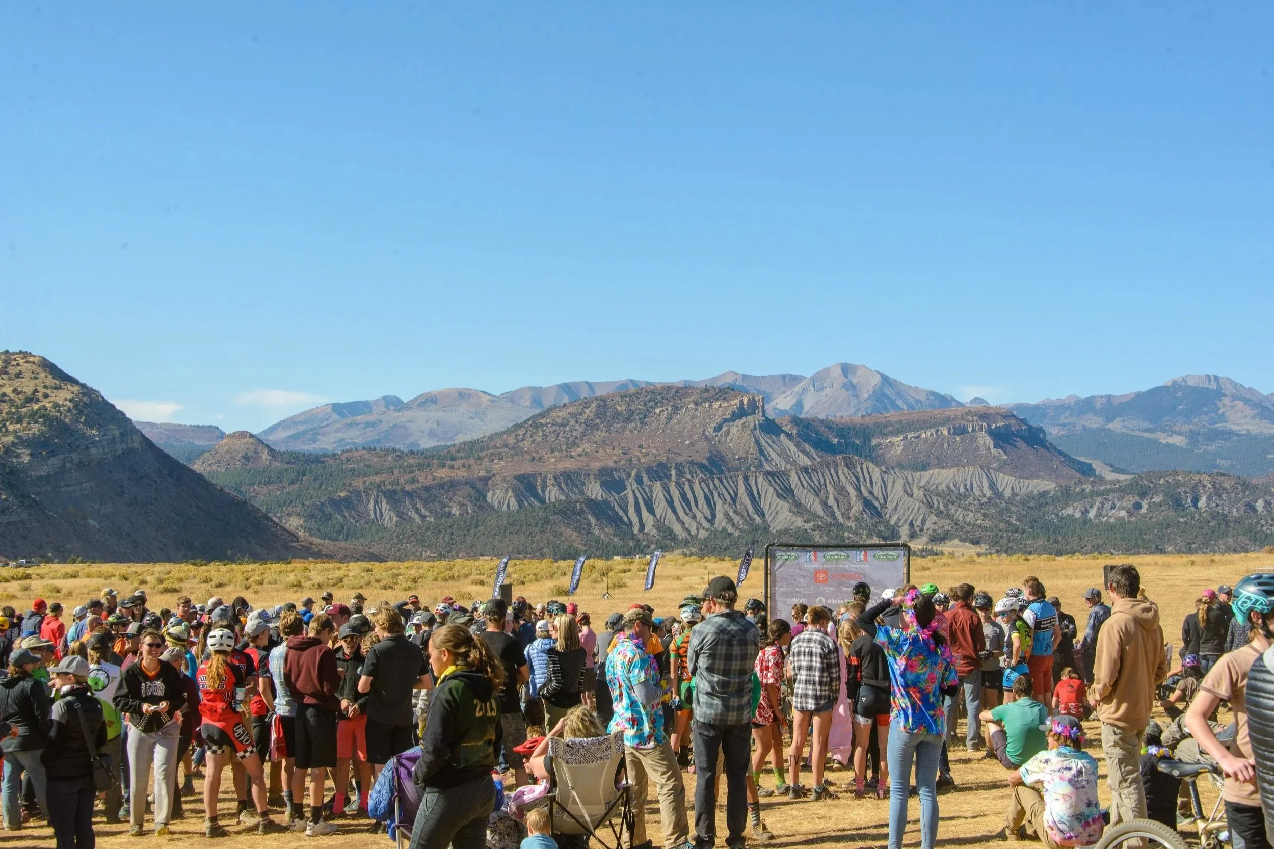 A large crowd of people gathered outdoors in front of a mountain landscape backdrop under a clear blue sky, some wearing helmets and casual clothing, possibly attending an event or race.