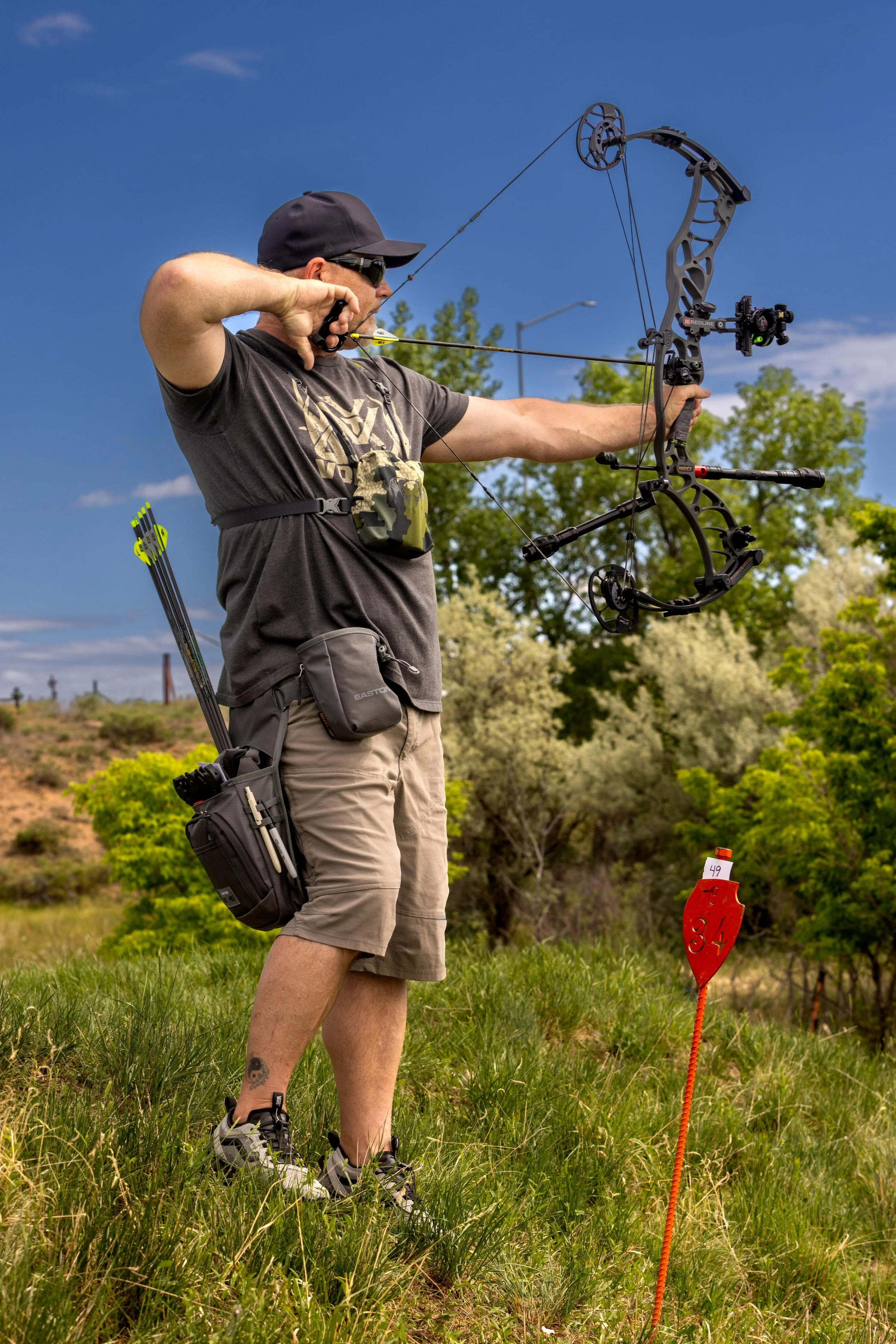 Man practicing archery outdoors, aiming with a compound bow, wearing a black baseball cap, sunglasses, a dark t-shirt, khaki shorts, and a tactical pouch, with a quiver of arrows on his side, green trees and a blue sky in the background.