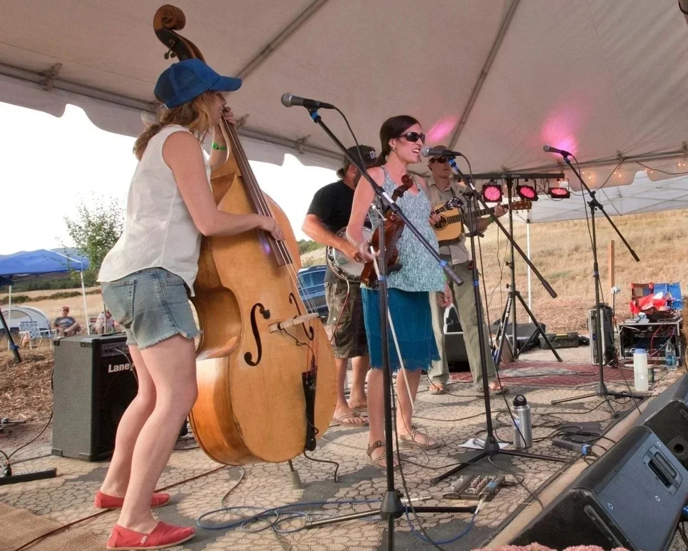 A band of five musicians performing outdoors under a tent. One woman is singing into a microphone while playing a violin, another woman is playing a double bass, and three men are playing guitars. There are various microphones, amps, and equipment on the stage, with a field and some people in the background.