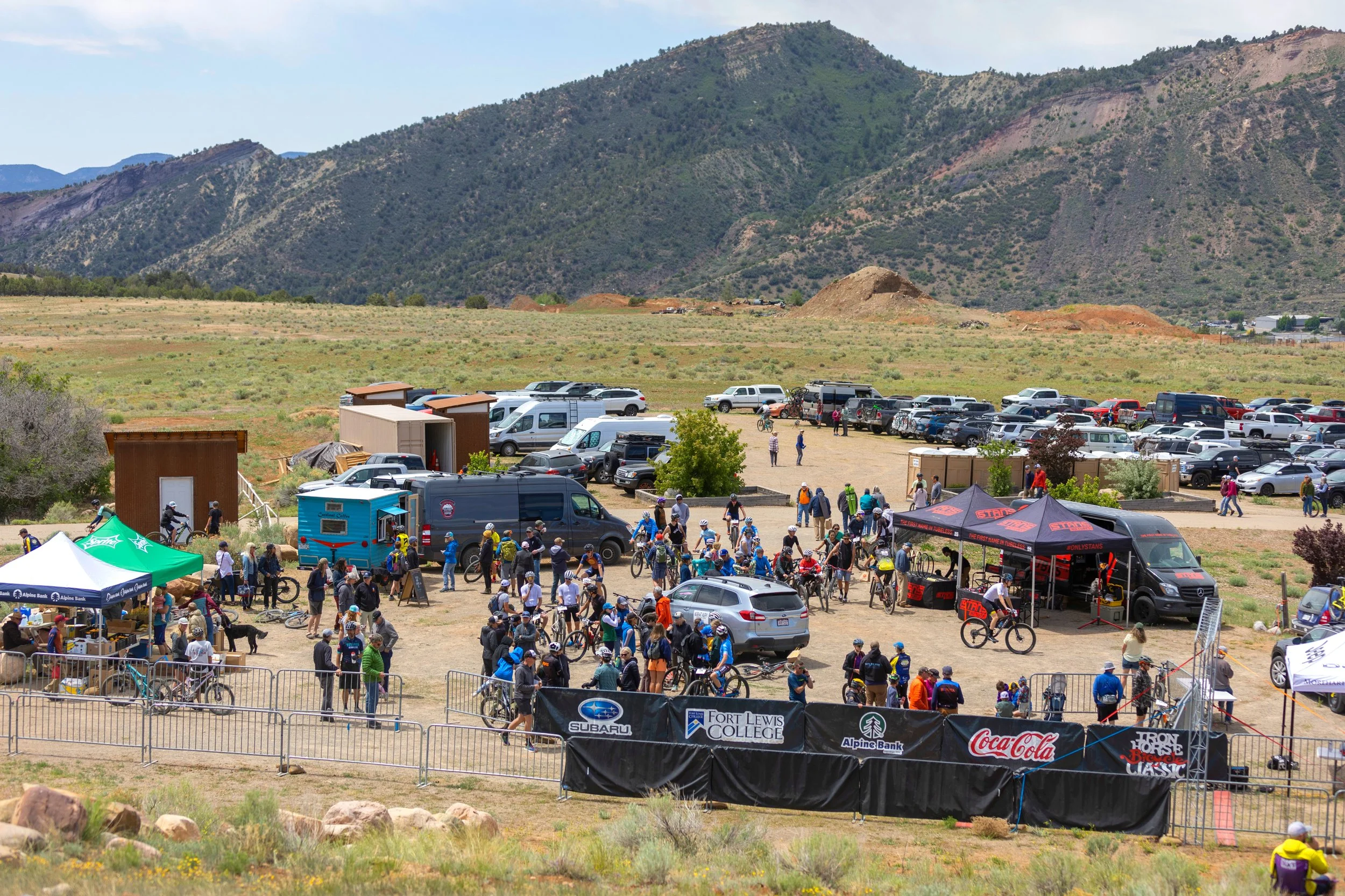 Crowd gathered at an outdoor event with tents, banners, and parked cars, set against a backdrop of mountains and cloudy sky.