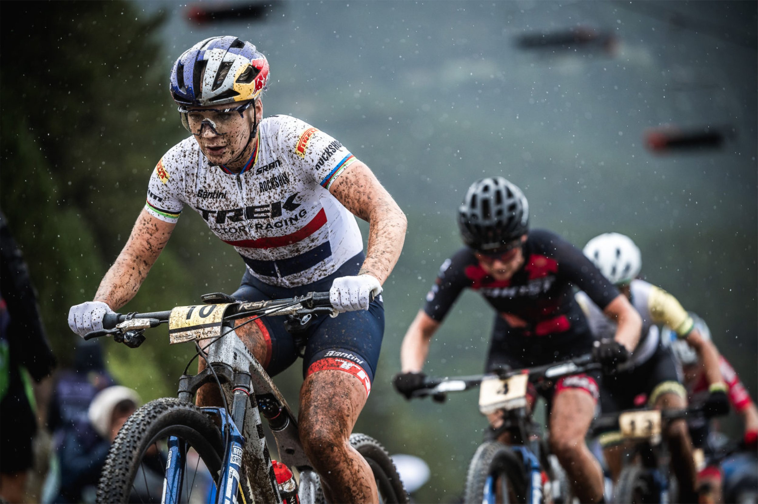 Cyclists participating in a muddy mountain bike race, wearing helmets and racing gear, with a muddy, wet trail in the background.