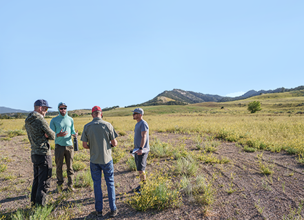 Five men standing and talking in an open field with mountains in the background.