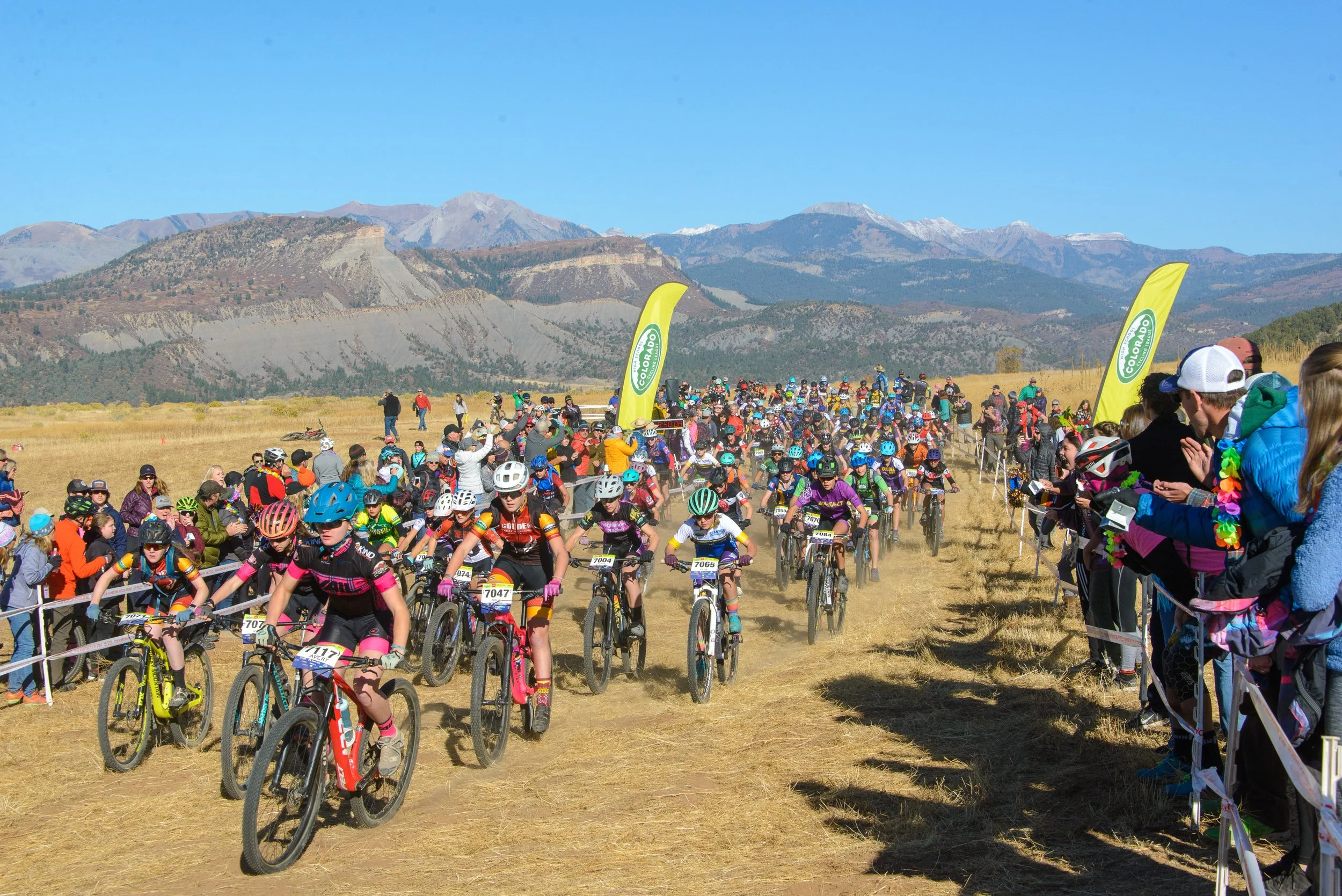 A large group of children and adults participating in a mountain biking event outdoors with mountains in the background. Many are wearing helmets, and some are taking photos or cheering.