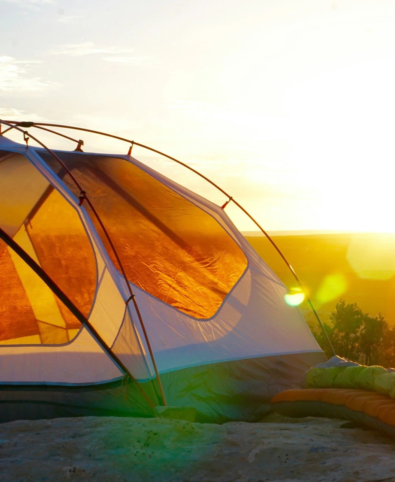 A camping tent set up on a sandy surface at sunset, with the sun shining brightly in the background and trees visible in the distance.