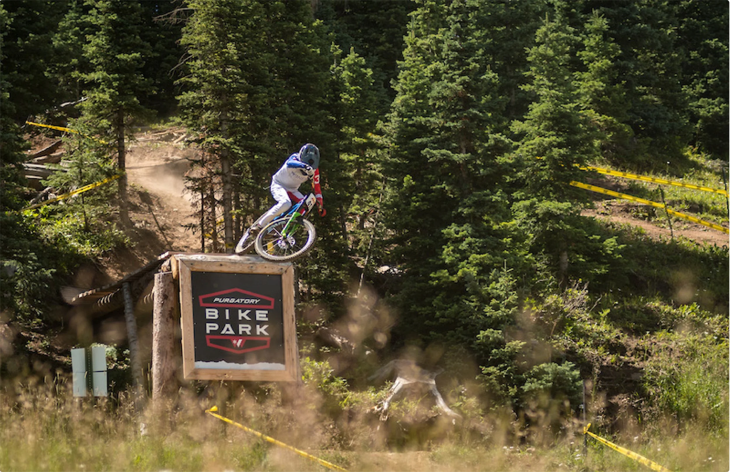A mountain biker wearing a helmet and gear riding down a dirt trail through a forested area at a bike park, with a sign reading 'Purgatory Bike Park' nearby.
