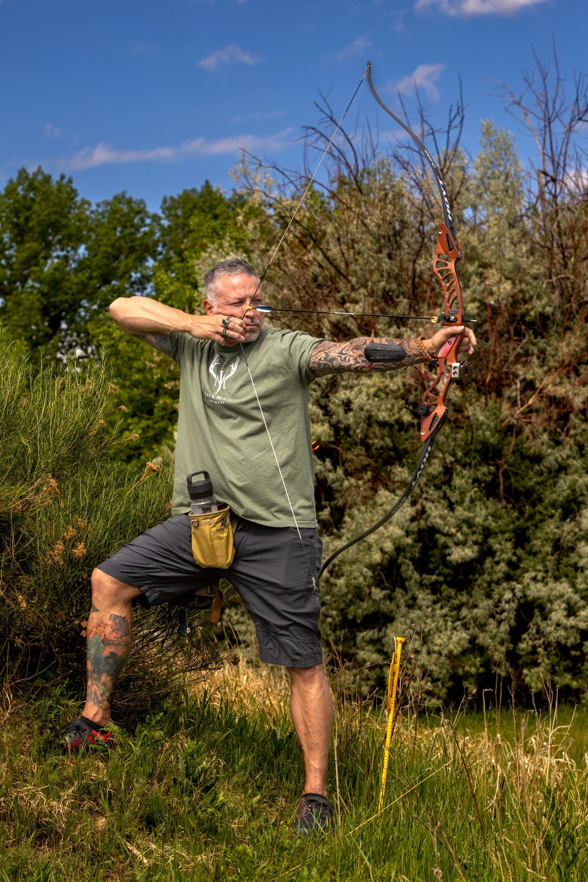Man dressed in green t-shirt and black shorts drawing a bow in a grassy outdoor setting with trees and a blue sky in the background.