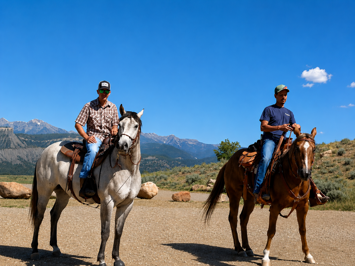 Two men riding horses in a mountainous landscape on a sunny day. One man is wearing a checkered shirt and sunglasses, on a gray horse. The other is wearing a blue shirt and cap, on a brown horse. There are rocks and green shrubs in the background with mountains and blue sky.