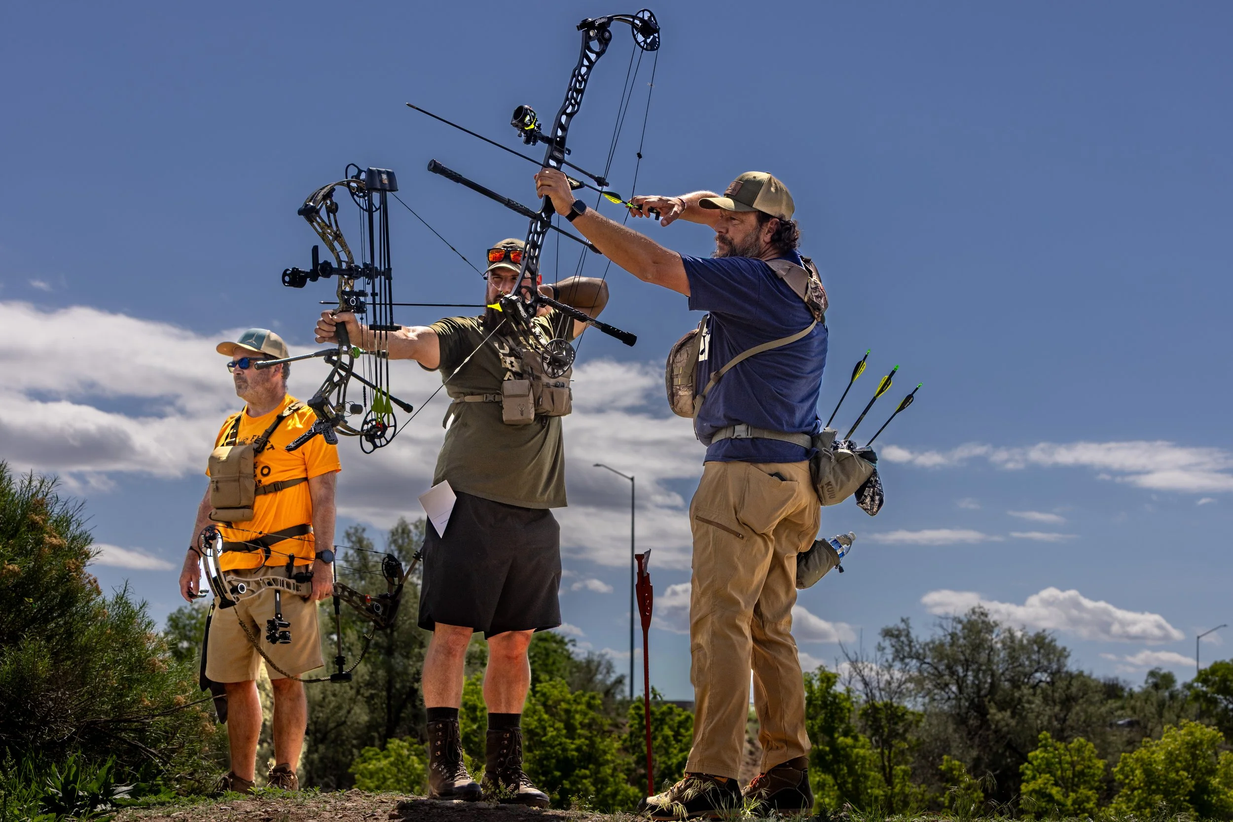 Three men on a grassy outdoor field, two holding bows and aiming, with a third man in the background, all dressed in outdoor gear, under a partly cloudy blue sky.