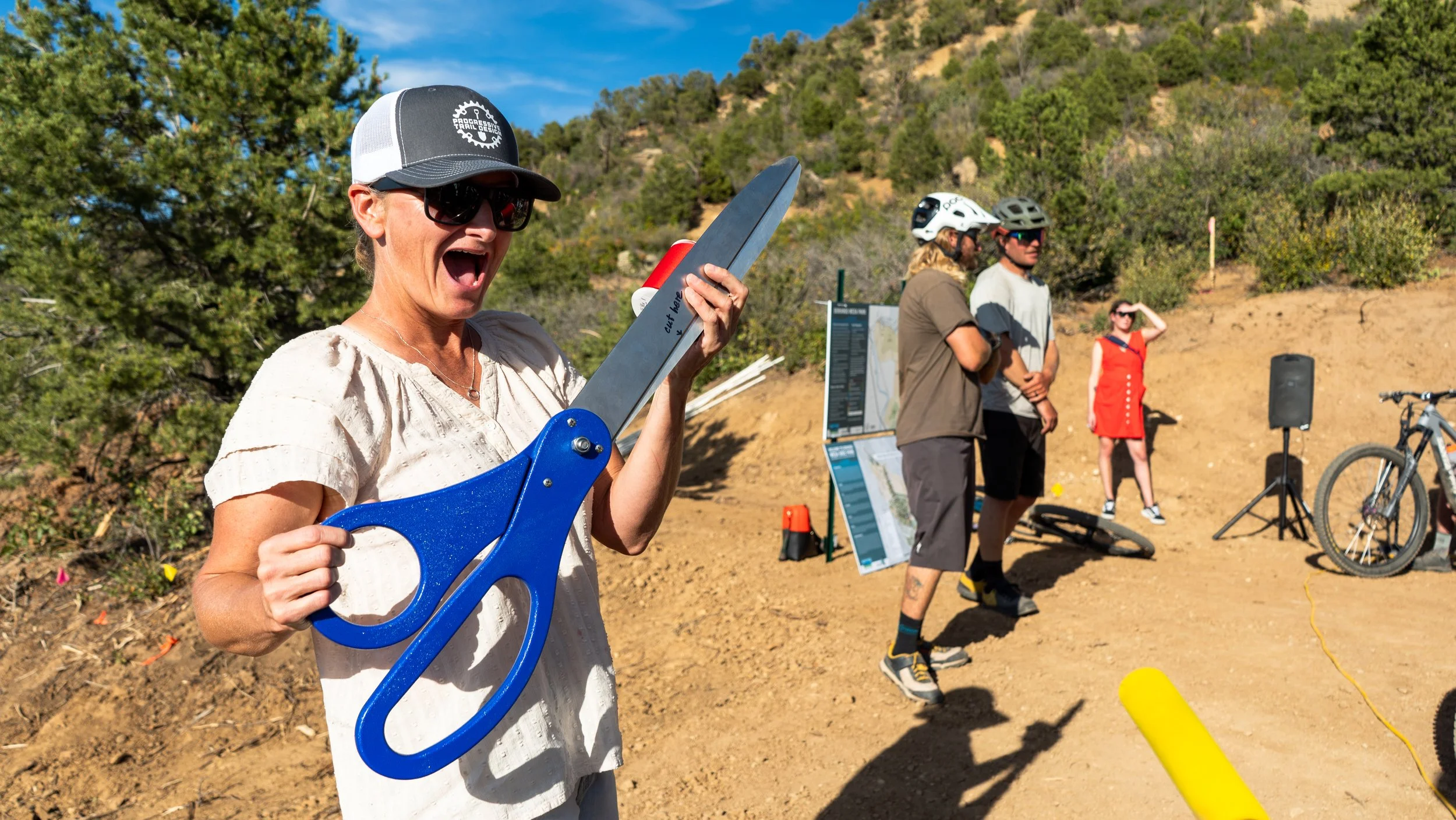 A woman wearing sunglasses and a cap is holding a large pair of novelty scissors, appearing to be about to cut a ribbon or cord. In the background, there are several people, some wearing helmets and casual outdoor clothing, standing outdoors on a dirt area with hills and trees behind them. A map or informational display and a bicycle are also visible.