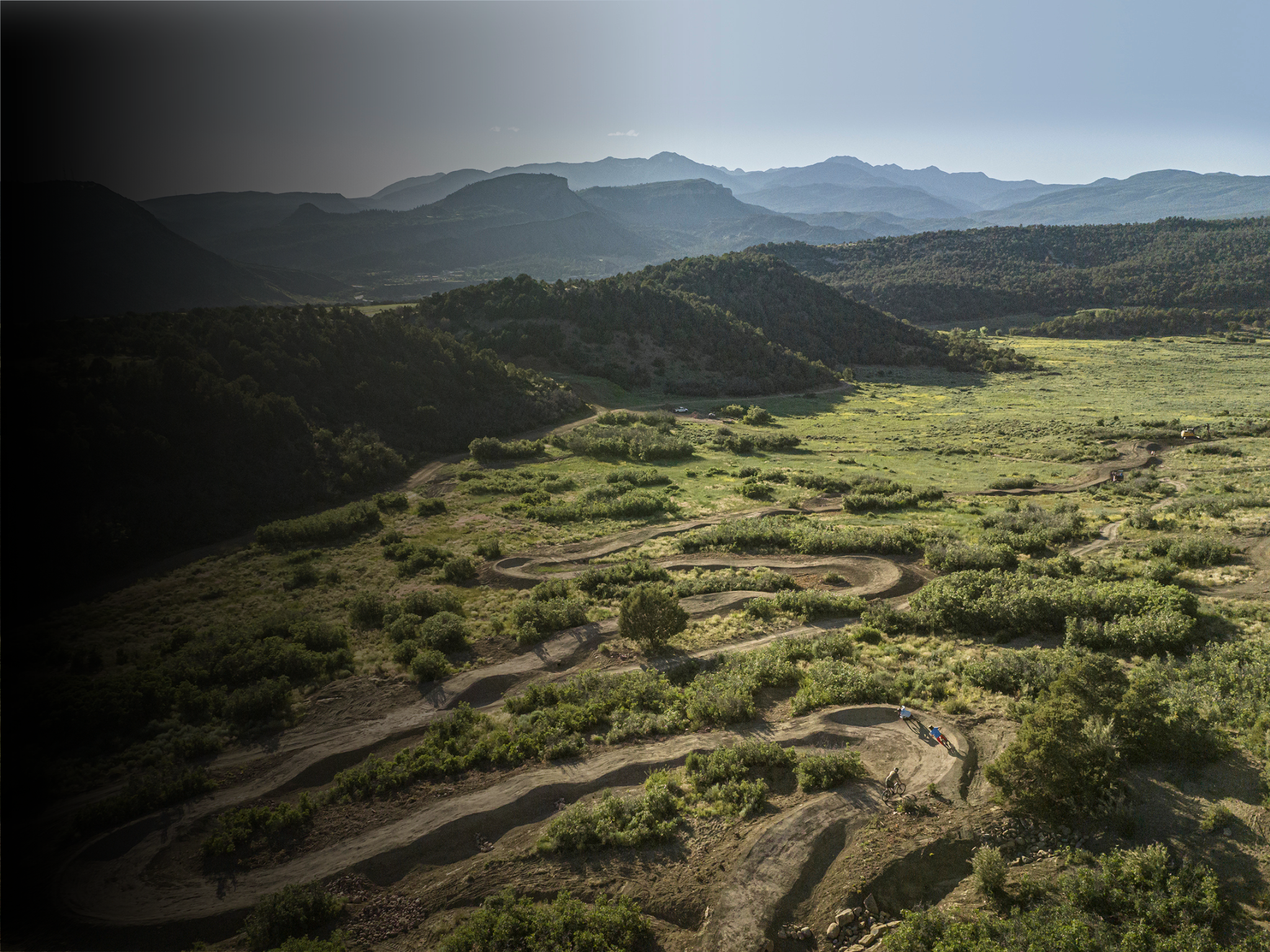 A scenic landscape of rolling green hills and mountains under a clear sky, with mountain bikers riding a dirt trail in the foreground.