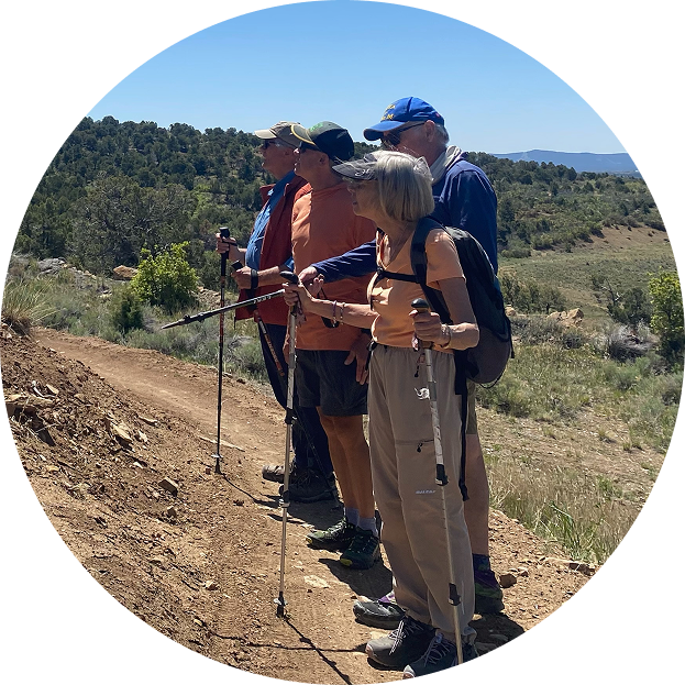 Five hikers standing on a dirt trail in a rural area, with a background of rolling hills and greenery, under a clear blue sky.