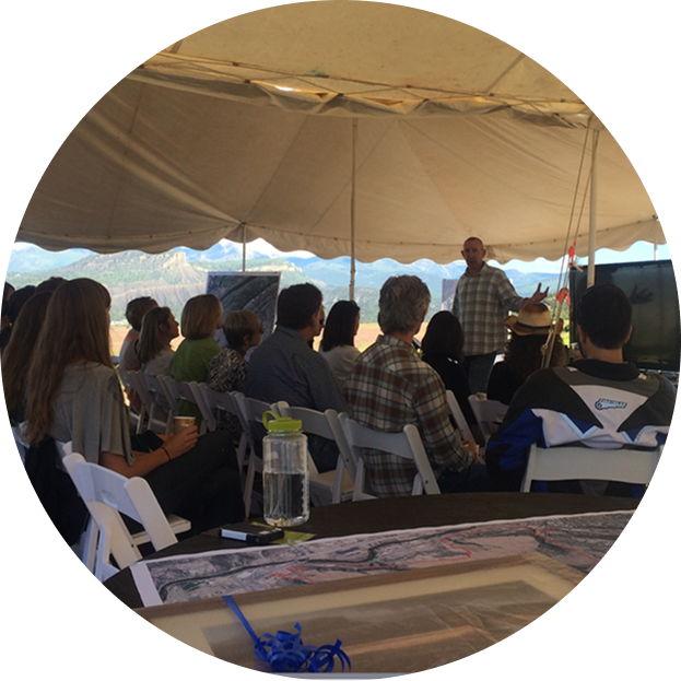 Group of people sitting under a large tent, listening to a man speaking at a presentation or meeting.