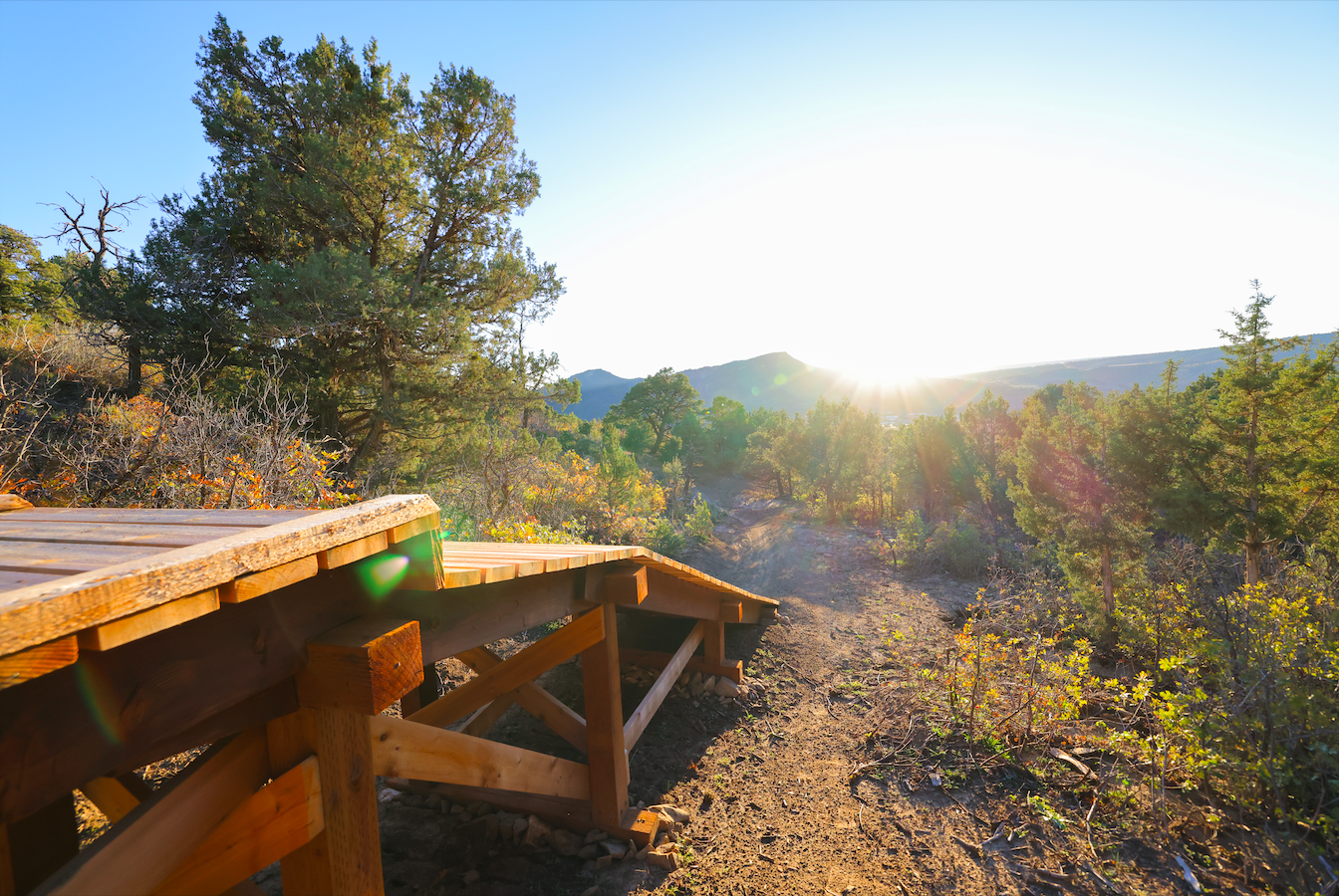 A wooden bridge on a trail in a forest with trees and mountains in the background, sunlight shining through as the sun sets or rises.