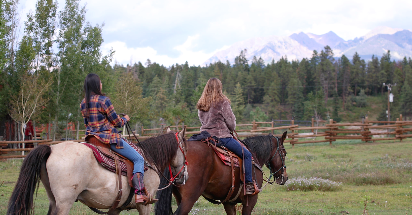 Two women riding horses in an open field with trees and mountains in the background.