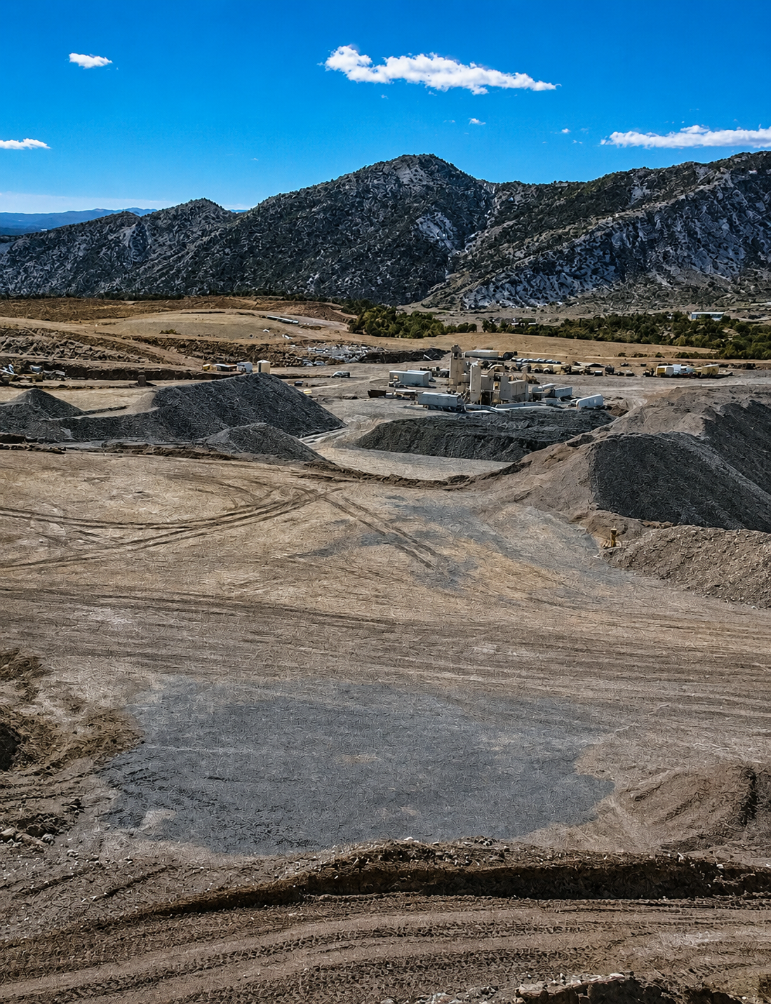 A landscape of a quarry with hills and mountains in the background, showing excavated earth and piles of dark rock, under a blue sky with some clouds.