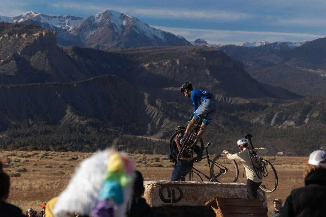 A skateboarder in a blue jersey performs a jump over a ramp while holding a bicycle, with two people nearby also holding bicycles, mountains and a blue sky in the background.