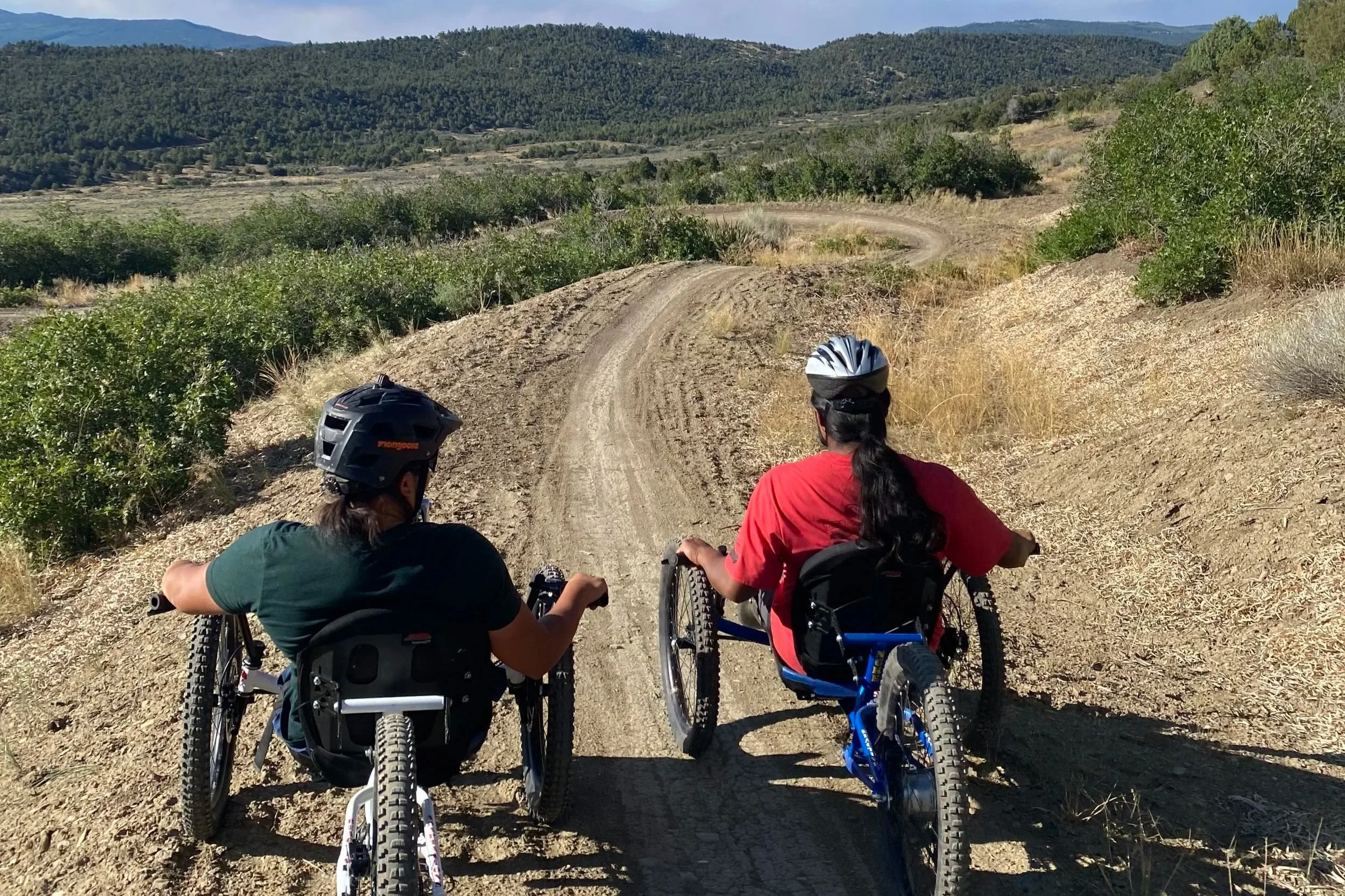 Three people riding mountain bikes on a dirt trail surrounded by green shrubs and trees, with hills and mountains in the background under a blue sky.