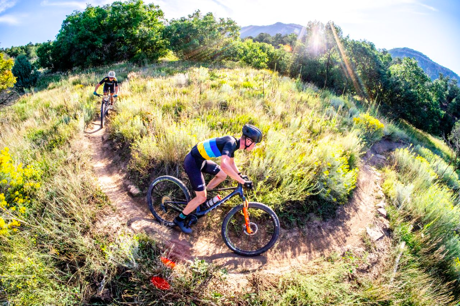 Two mountain bikers riding along a dirt trail on a sunny day in a lush, green, hilly area with mountains in the background.