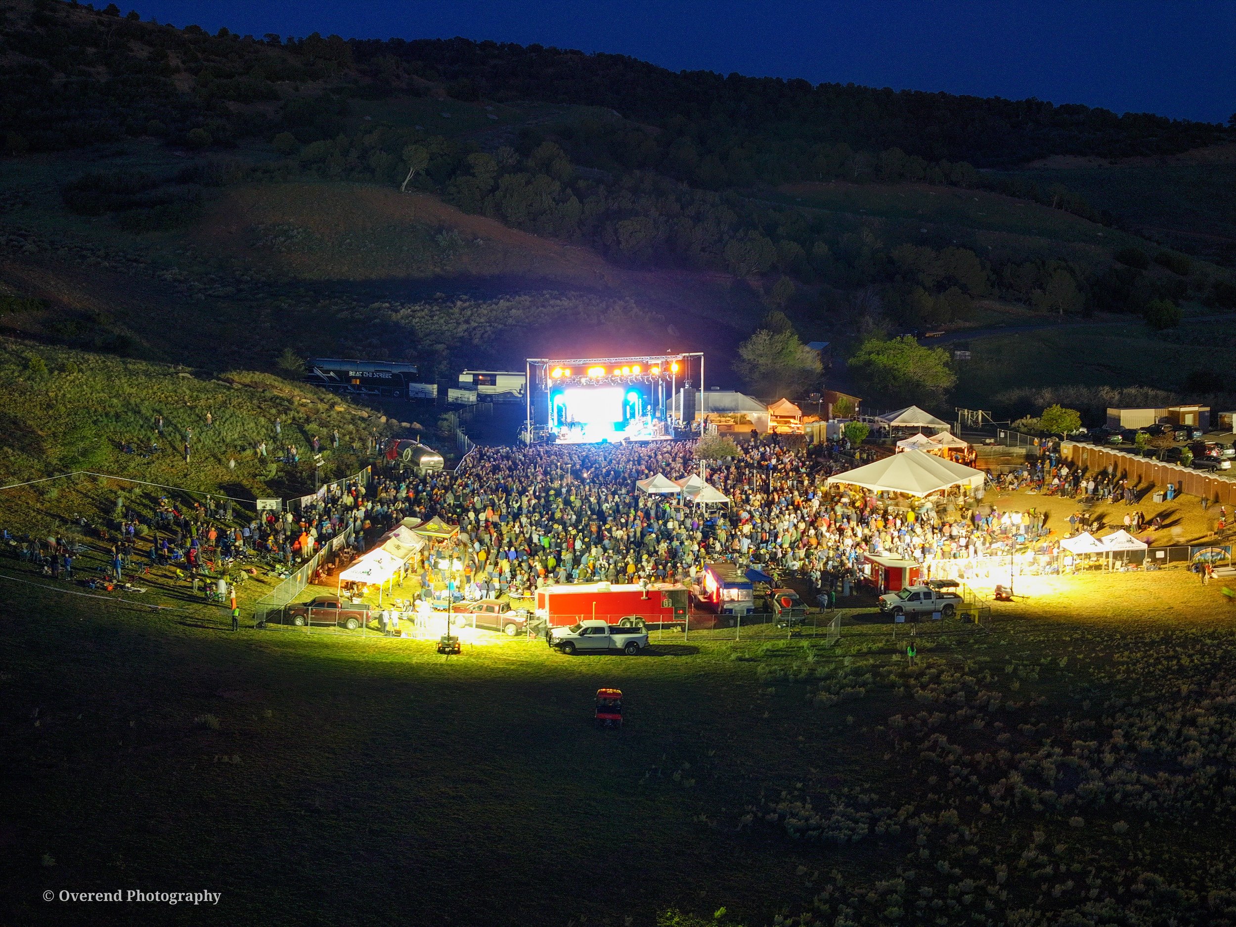 Nighttime outdoor concert with a large crowd in front of a brightly lit stage, set against dark hills and a blue sky.