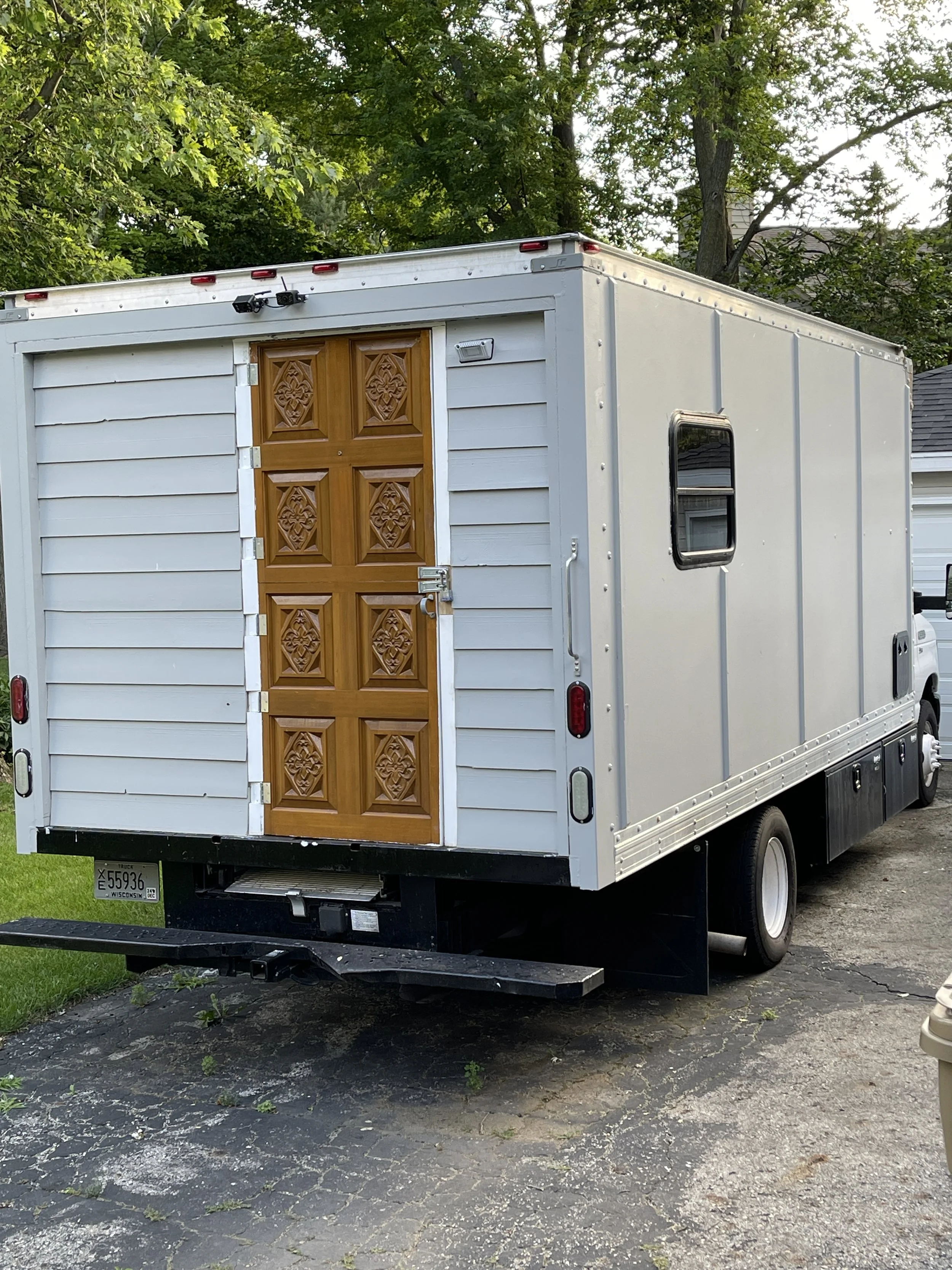 White mobile food trailer with a decorative wooden door, small side window, and a hitch for towing, parked on a paved driveway with green trees in the background.