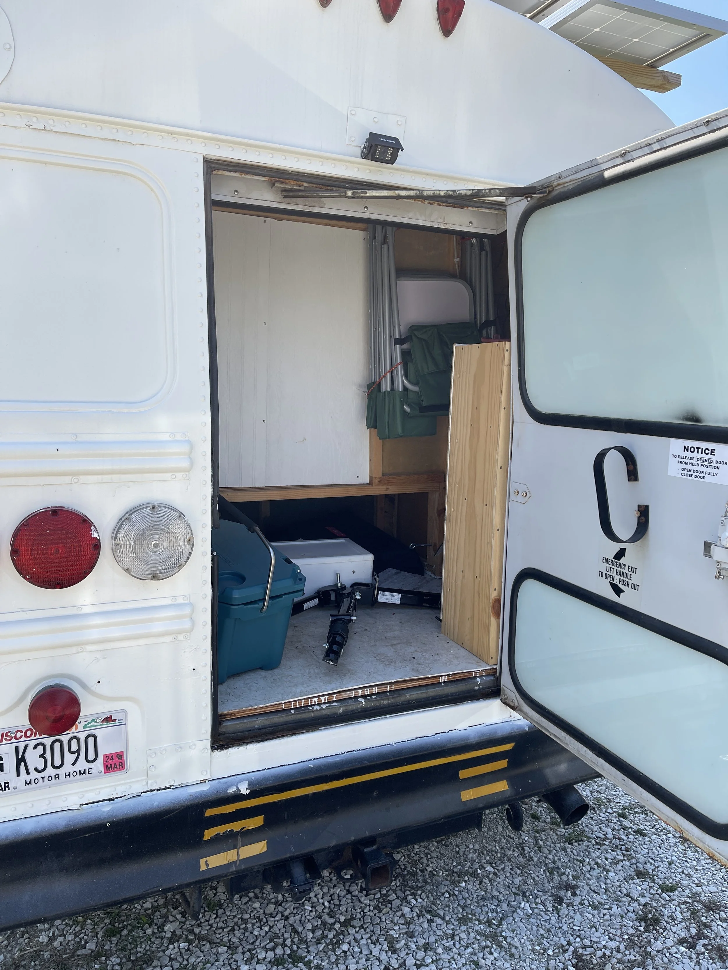 The open door of a converted school bus showing a small interior with a foldable chair, a storage box, and miscellaneous camping items visible.