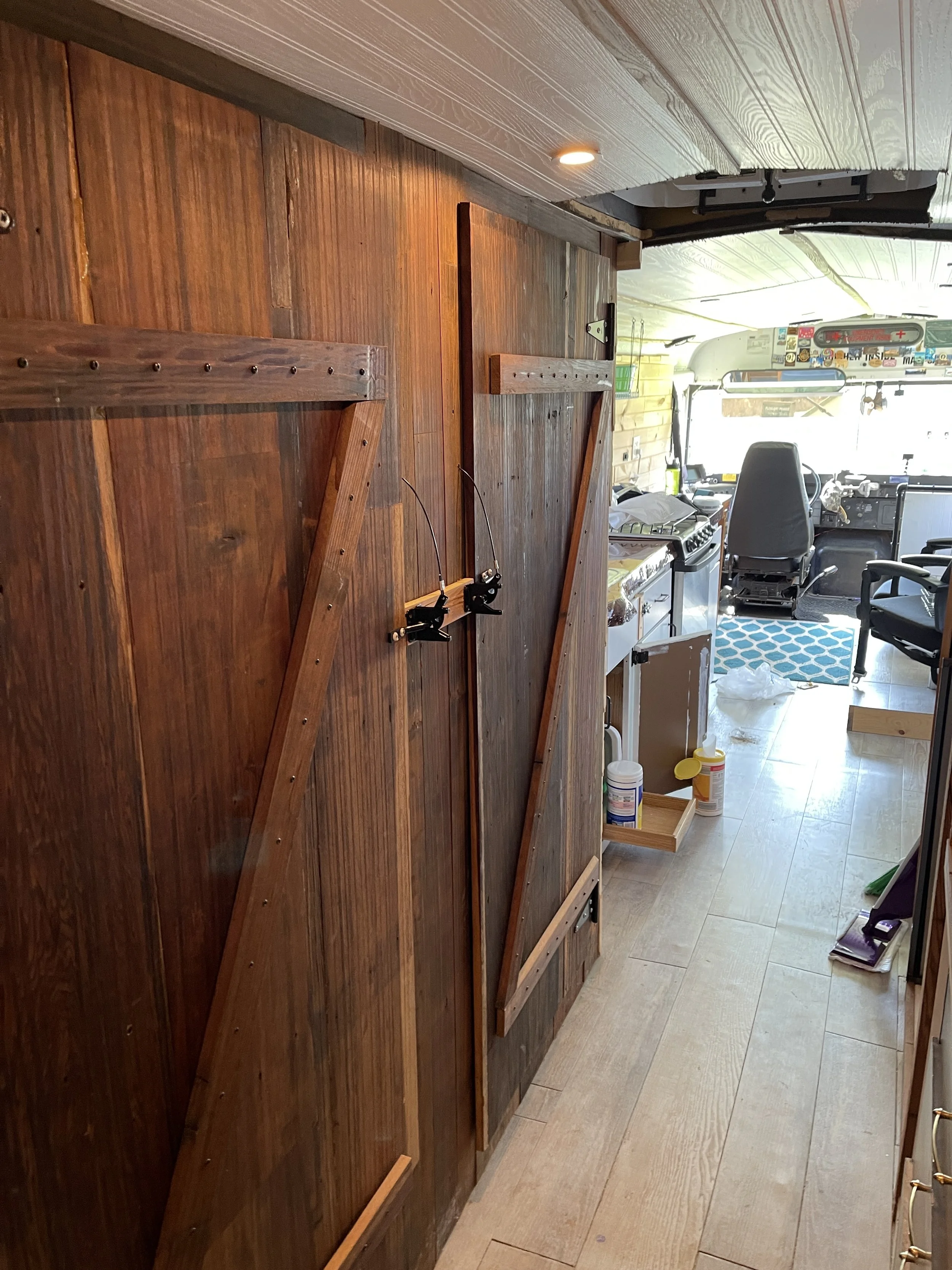 Interior of a camper van with a wooden wall, two black fishing rod holders, a small kitchen area with shelves and containers, and the driver's area with multiple stickers and a blue patterned mat on the floor.